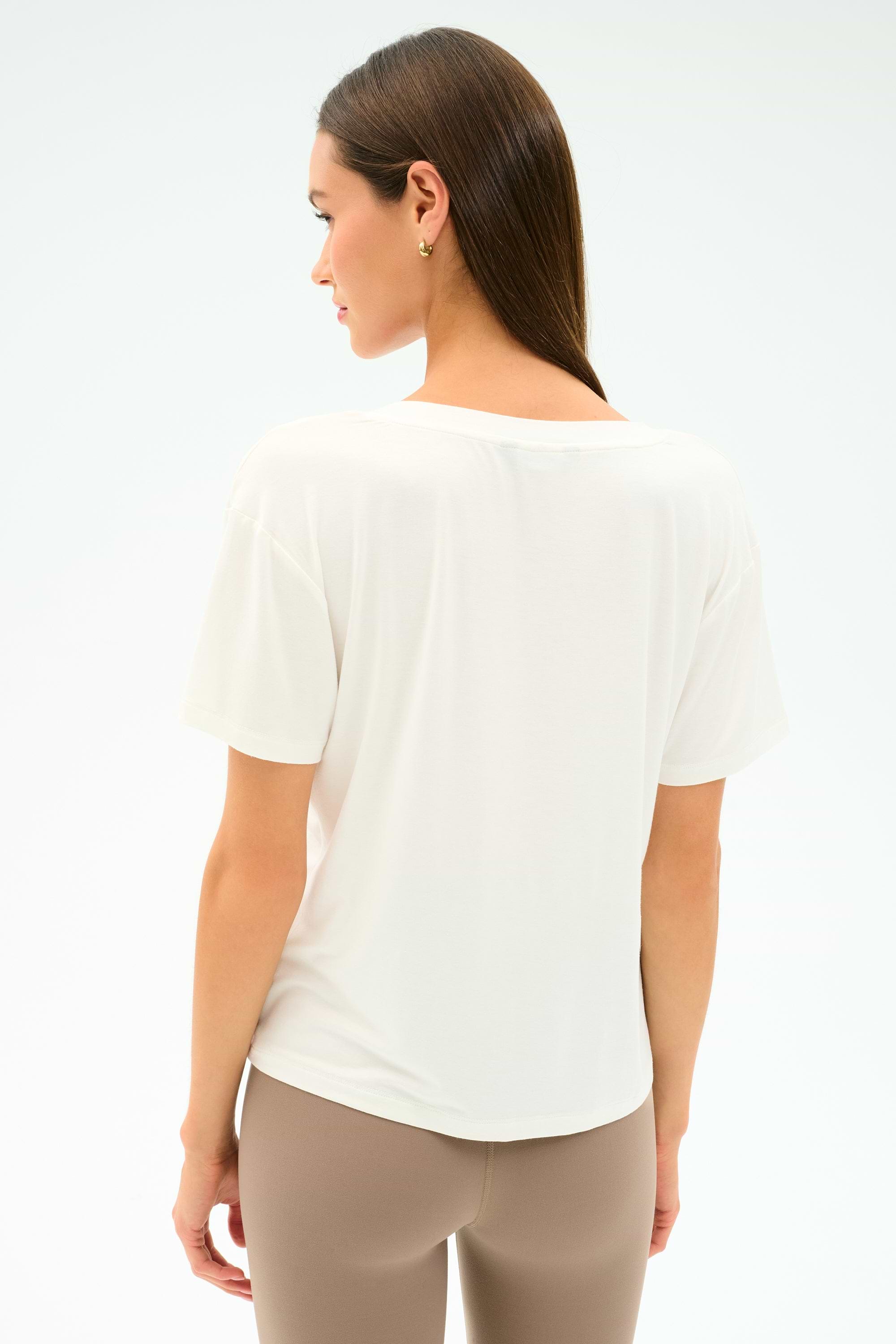 A woman stands with her back to the camera, wearing the SPLITS59 Georgia Jersey V-Neck in white and beige leggings, made from a soft viscose-spandex blend, set against a plain light background.