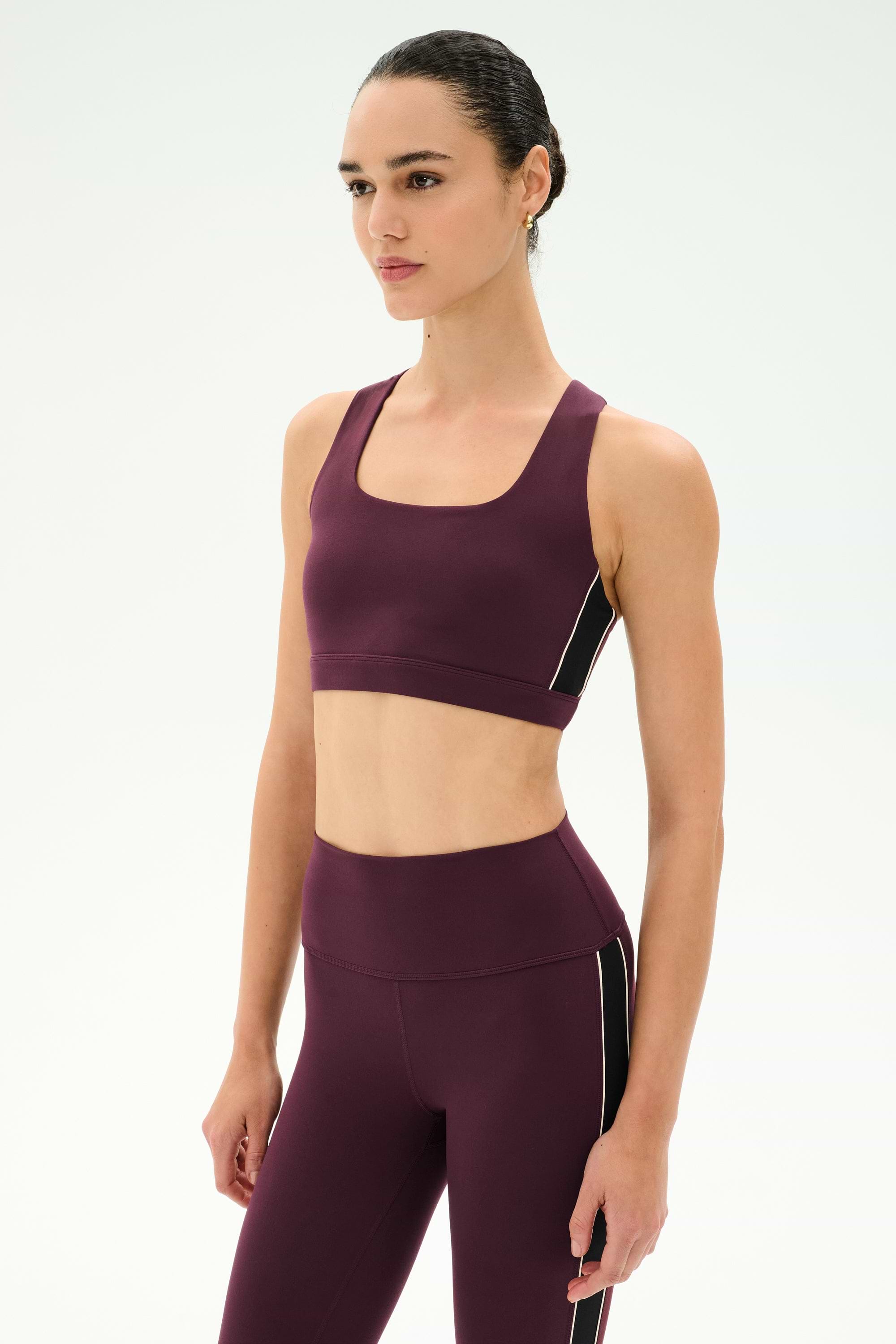 A person wearing the SPLITS59 Lexi Rigor Bra in Dark Cherry/Black, paired with matching leggings made from soft spandex and nylon, stands against a plain white background and gazes slightly to the side.