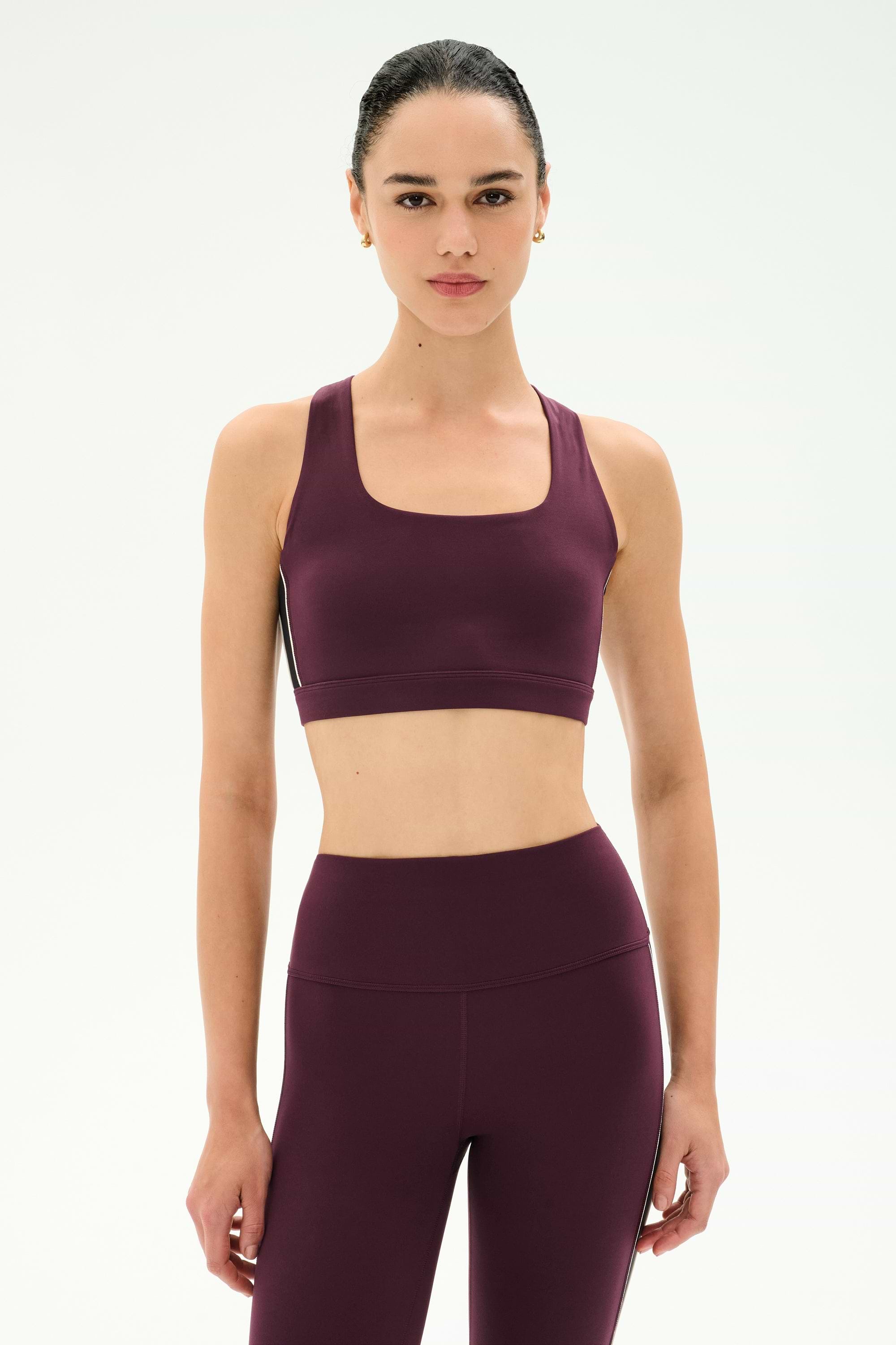 A woman stands against a plain light background wearing the SPLITS59 Lexi Rigor Bra in Dark Cherry/Black, paired with matching high-waisted leggings.