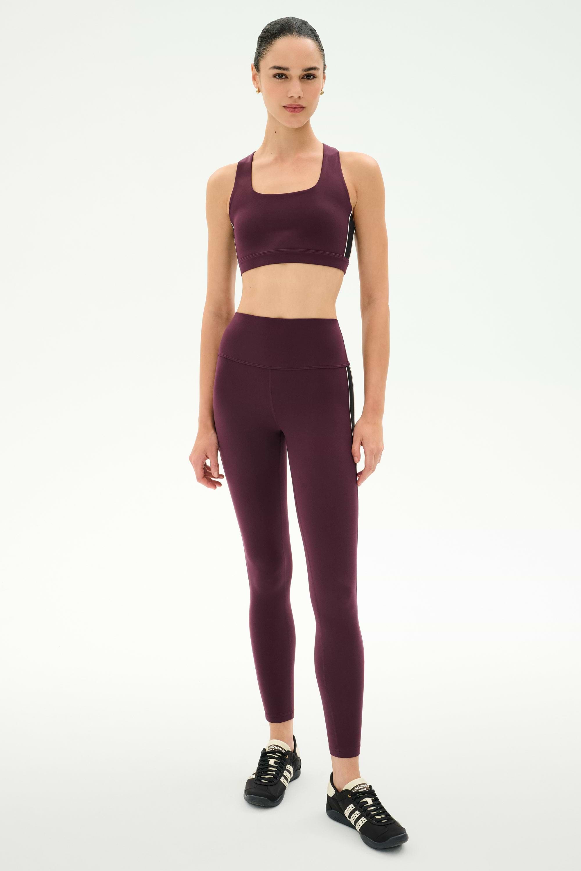 A woman stands in a studio against a plain white background, wearing the SPLITS59 Lexi Rigor Bra in Dark Cherry/Black paired with matching leggings and black sneakers.