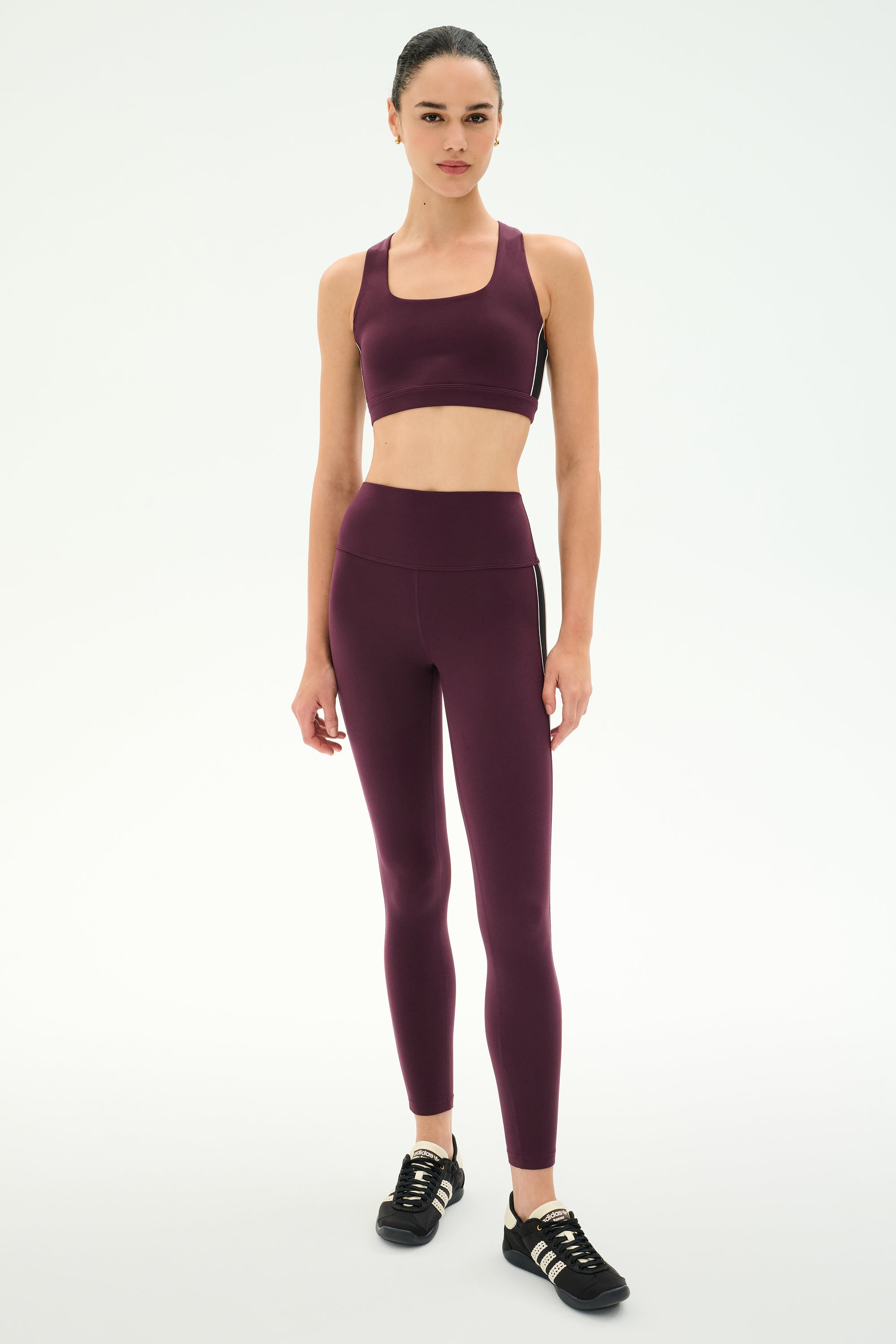 A woman stands against a plain background wearing the SPLITS59 Margot Rigor Legging in Dark Cherry/Black, paired with a matching sports bra, designed for high-performance gym workouts, and black athletic shoes.