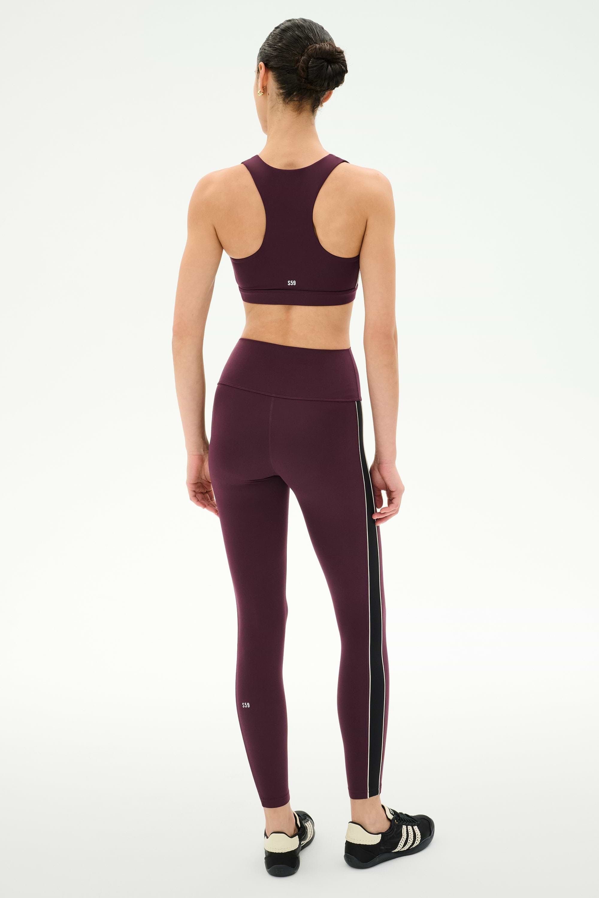 A person stands facing away, wearing the SPLITS59 Lexi Rigor Bra in Dark Cherry/Black paired with matching leggings made from a soft nylon-spandex blend featuring white side stripes, and black athletic shoes, set against a plain white background.