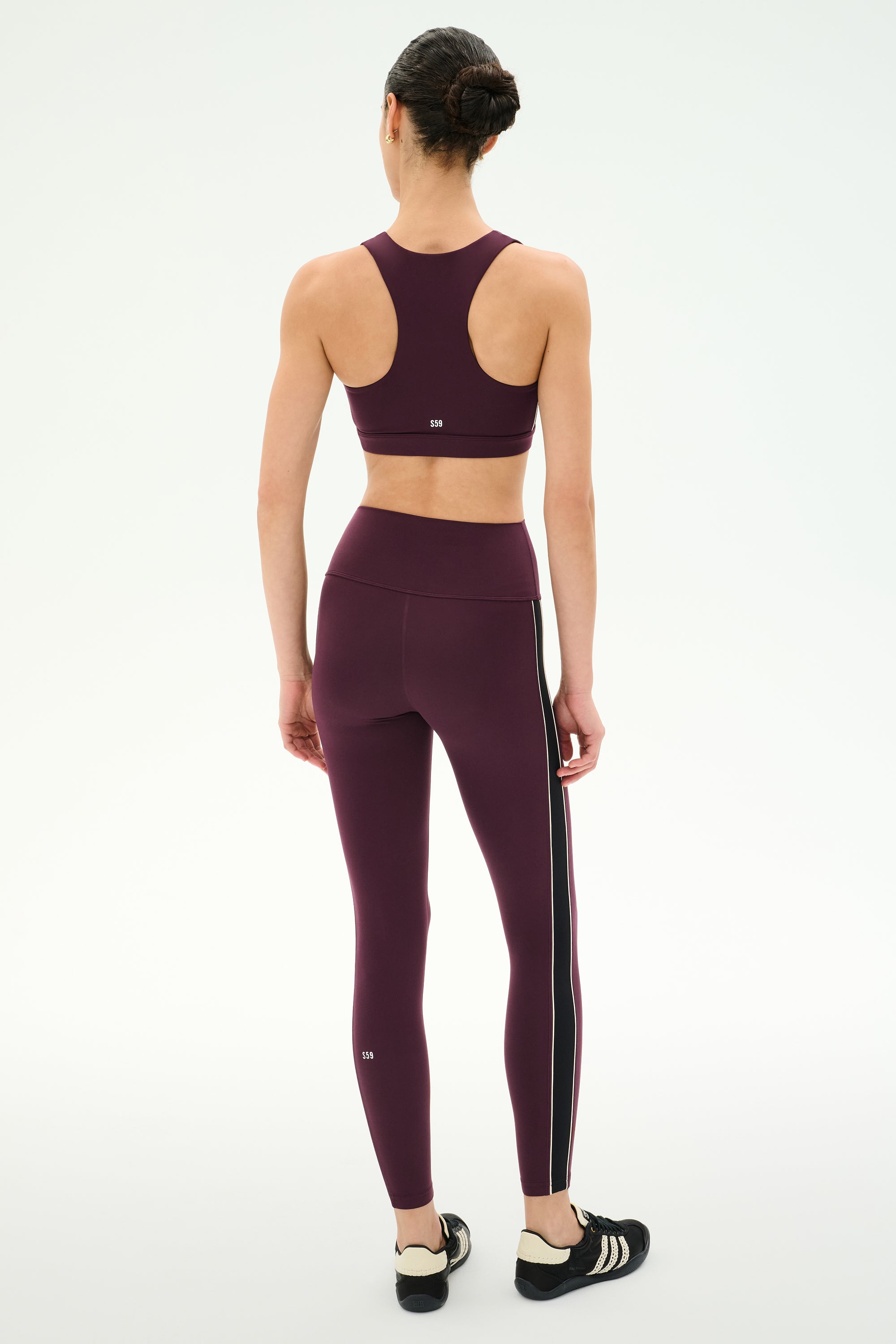 A person stands facing away, wearing the SPLITS59 Margot Rigor Legging in Dark Cherry/Black paired with a matching sports bra and black athletic shoes, perfect for gym workouts, against a plain white background.