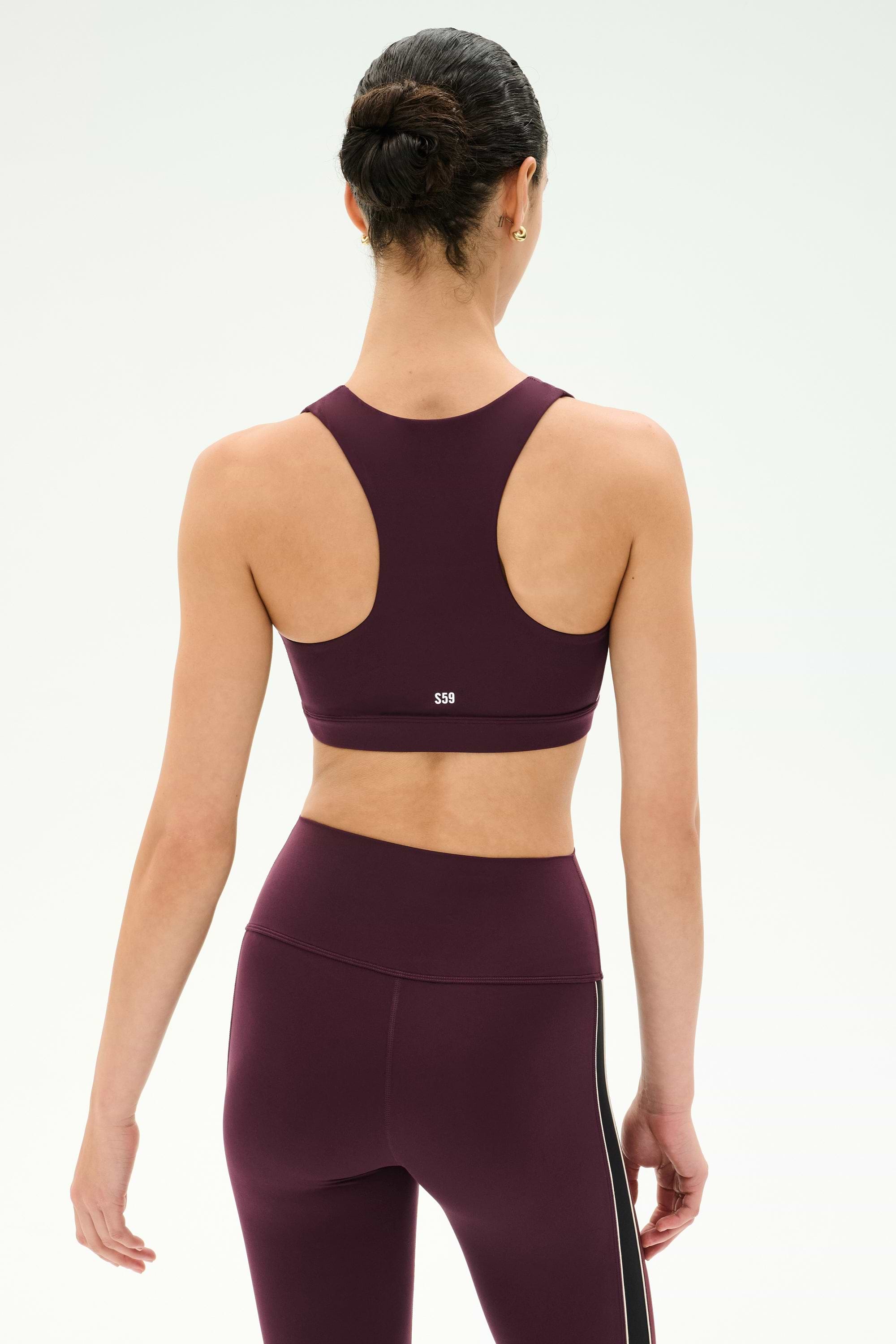A woman with dark hair in a bun is shown from the back against a plain background, wearing the SPLITS59 Lexi Rigor Bra in Dark Cherry/Black, paired with matching leggings.