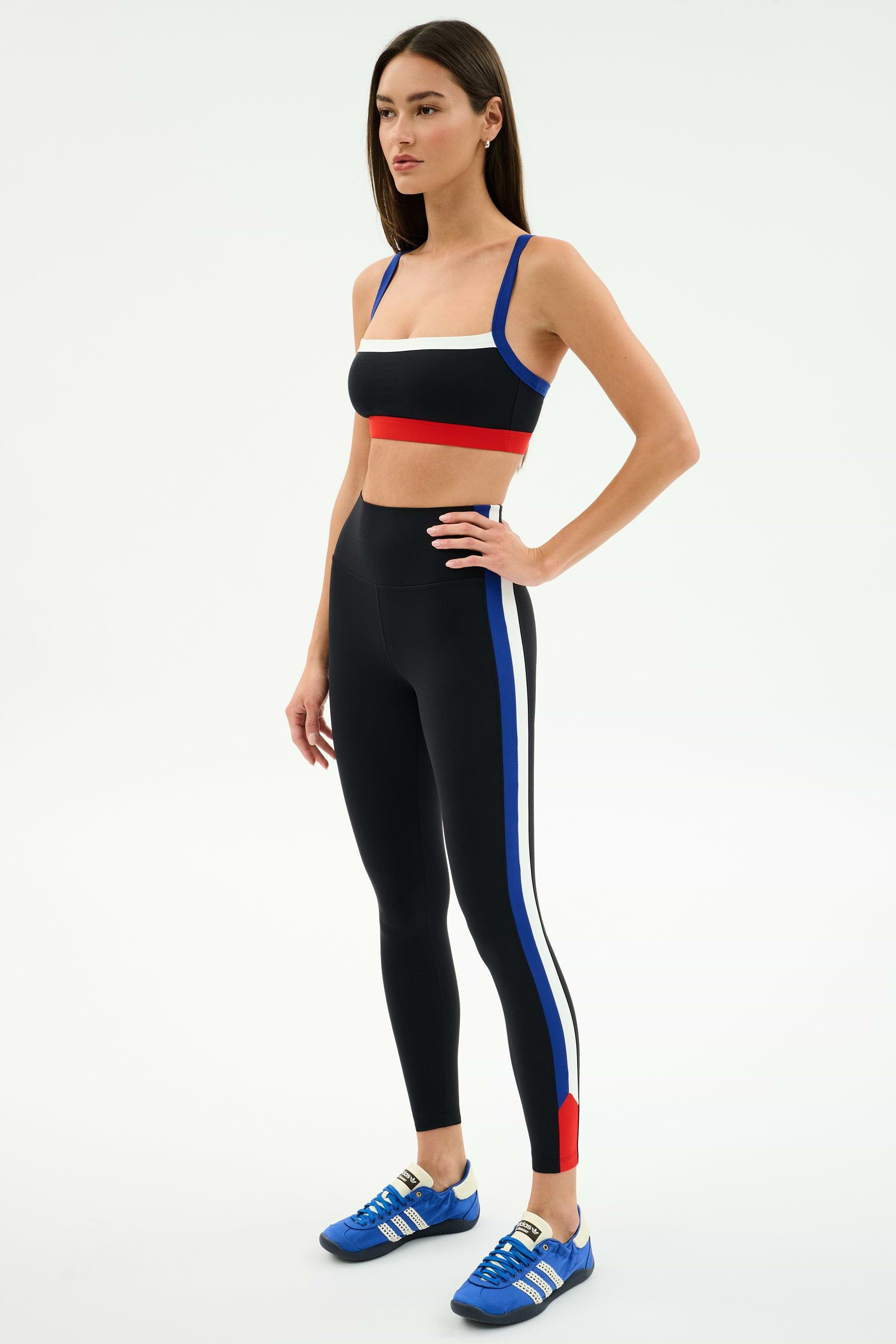 A woman wears SPLITS59's Monah High Waist Rigor Legging 7/8 in Black/Cobalt with blue, white, and red stripes and a matching black sports bra, paired with blue Adidas sneakers against a plain white background.