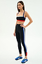 A woman wears SPLITS59's Monah High Waist Rigor Legging 7/8 in Black/Cobalt with blue, white, and red stripes and a matching black sports bra, paired with blue Adidas sneakers against a plain white background.