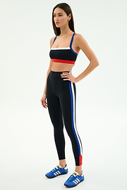 Woman wearing the SPLITS59 Monah Rigor Bra in Black/Cobalt and matching leggings with blue, white, and red stripes, paired with blue athletic shoes, stands against a plain white background.