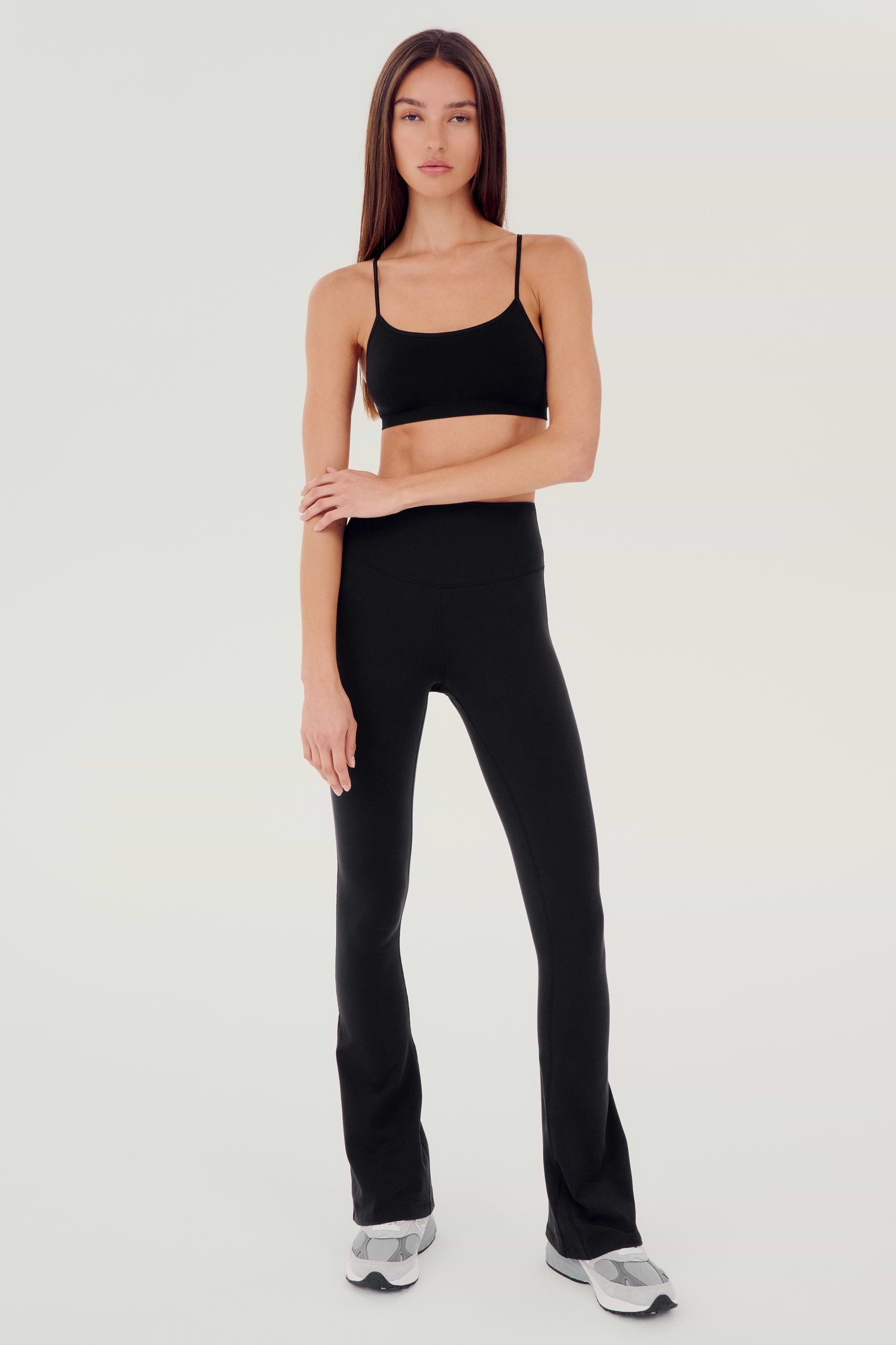 Full front view of woman with dark straight hair wearing black high waist below ankle length legging with wide flared bottoms and black racerback bra with spaghetti straps. Paired with white and gray shoes.
