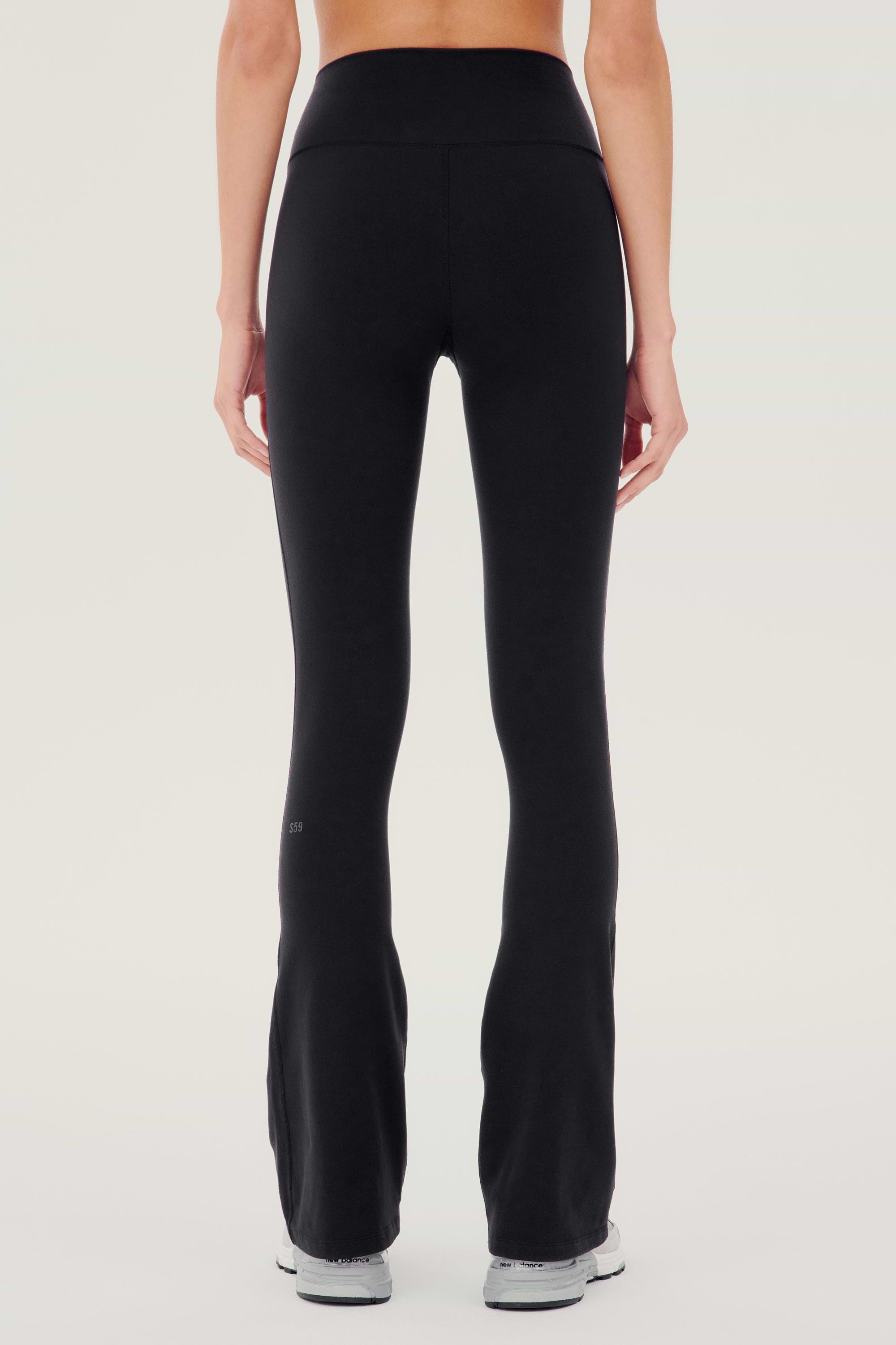 Back side view of woman wearing black high waist below ankle length legging with wide flared bottoms and gray S59 logo on bottom left calf. Paired with white and gray shoes.