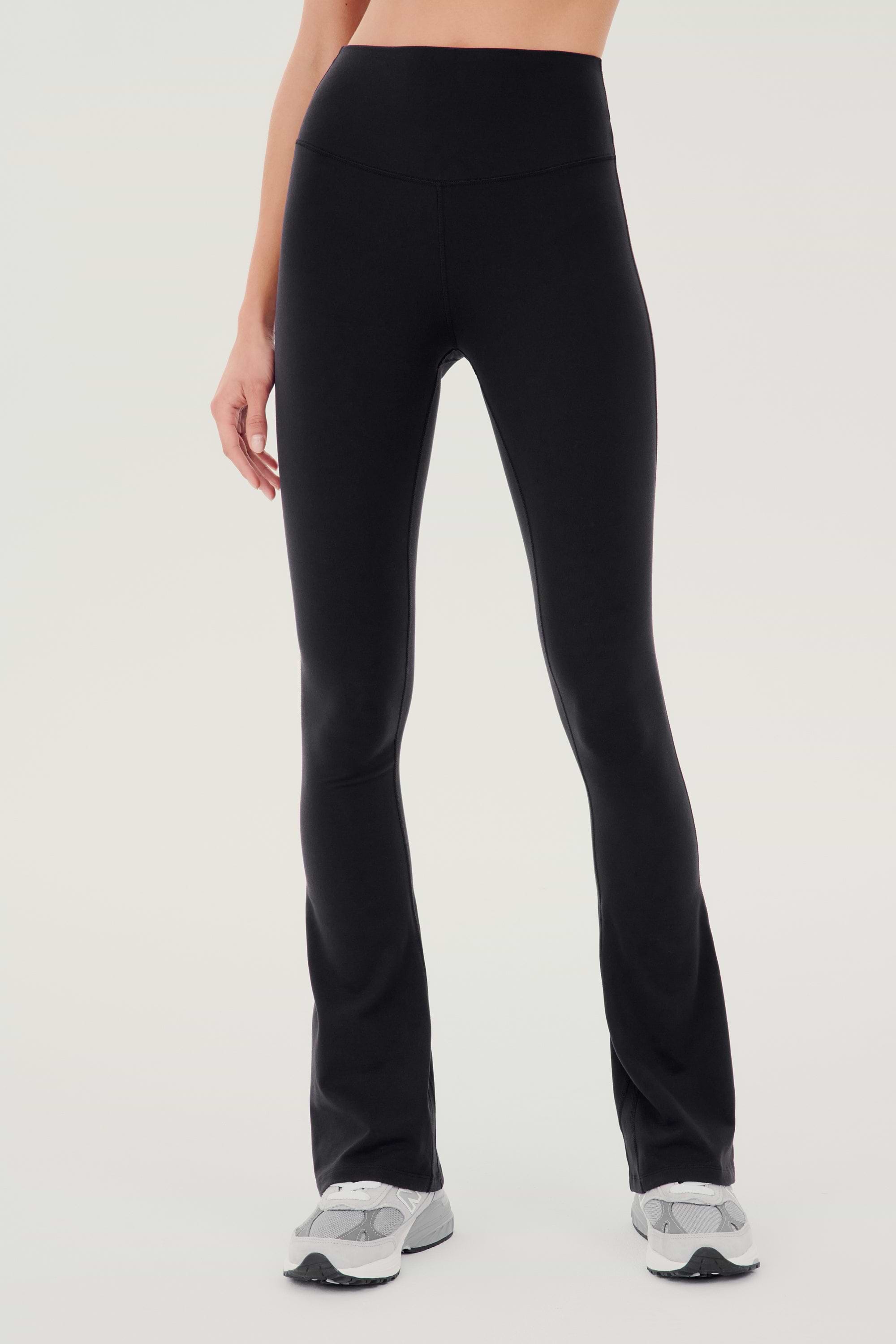 Front view of woman wearing black high waist below ankle length legging with wide flared bottoms. Paired with white and gray shoes.