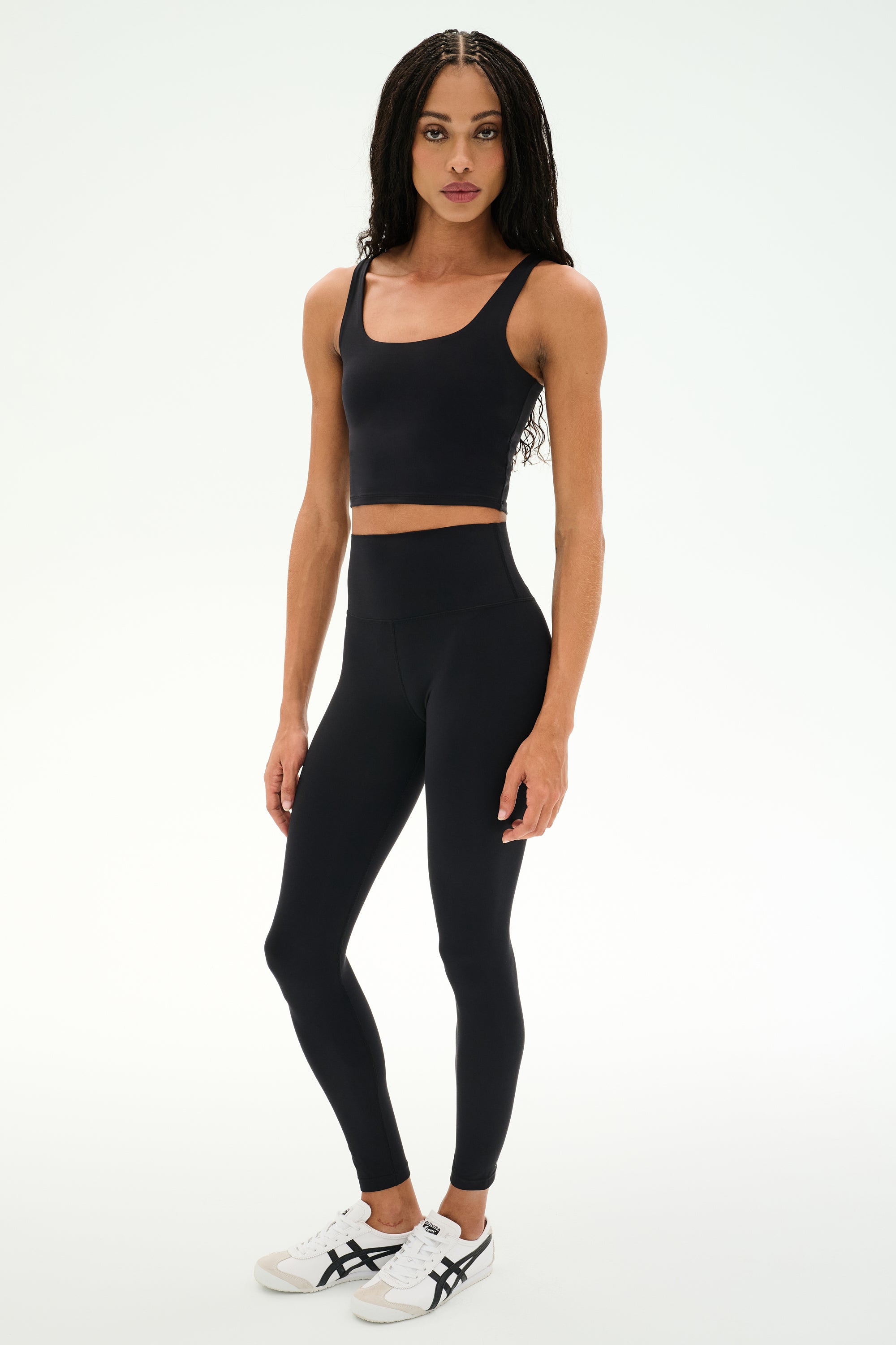 A person stands against a white background, wearing a black crop top, SPLITS59 Sprint High Waist Rigor 7/8 leggings in black, and white sneakers with black stripes.