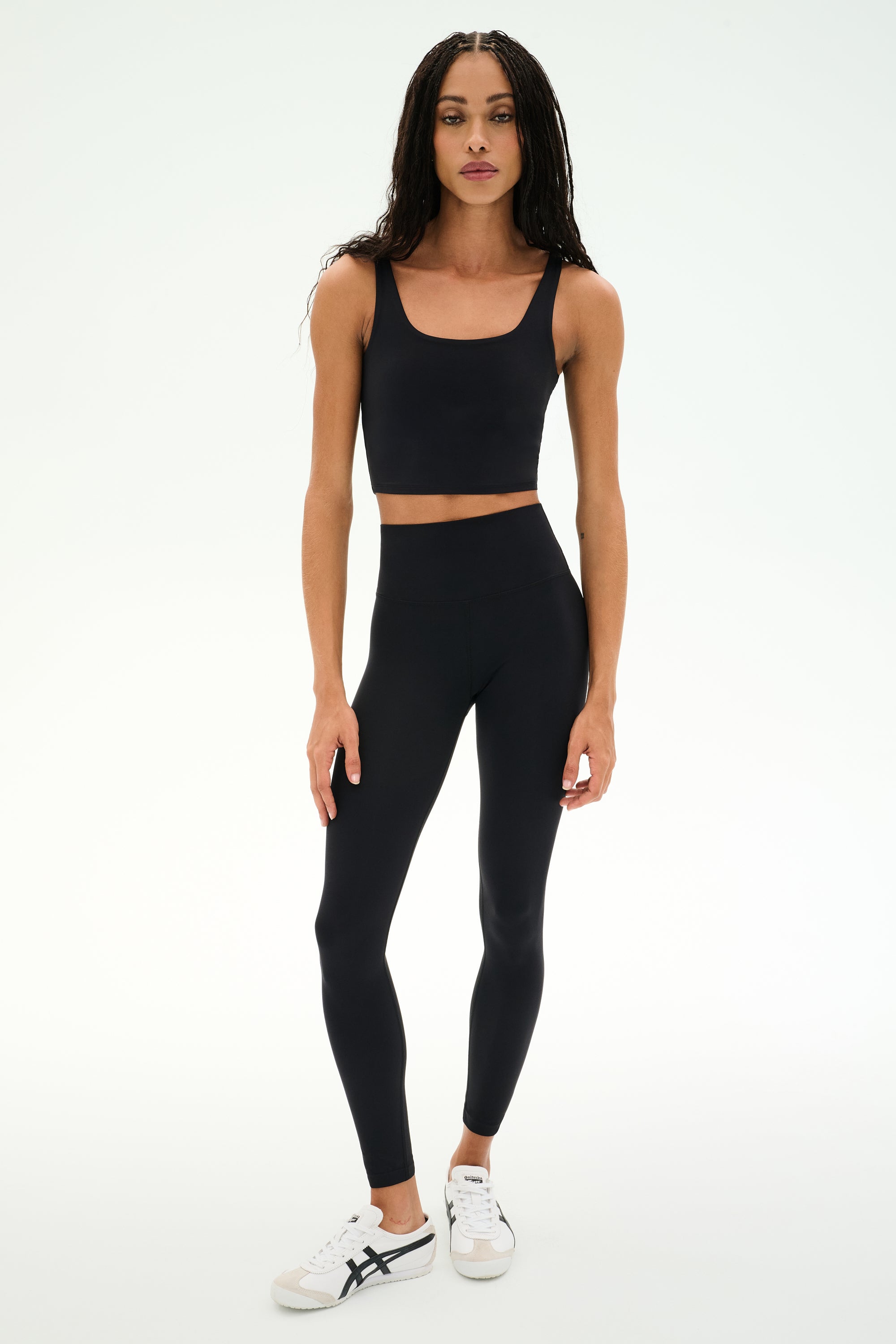 A woman stands against a white background, wearing a black crop top, white sneakers, and the SPLITS59 Sprint High Waist Rigor 7/8 leggings in black—crafted from durable Rigor fabric.