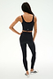 A person stands facing away against a plain white background, wearing SPLITS59’s Sprint High Waist Rigor 7/8 leggings in black with a matching black athletic crop top and white sneakers.