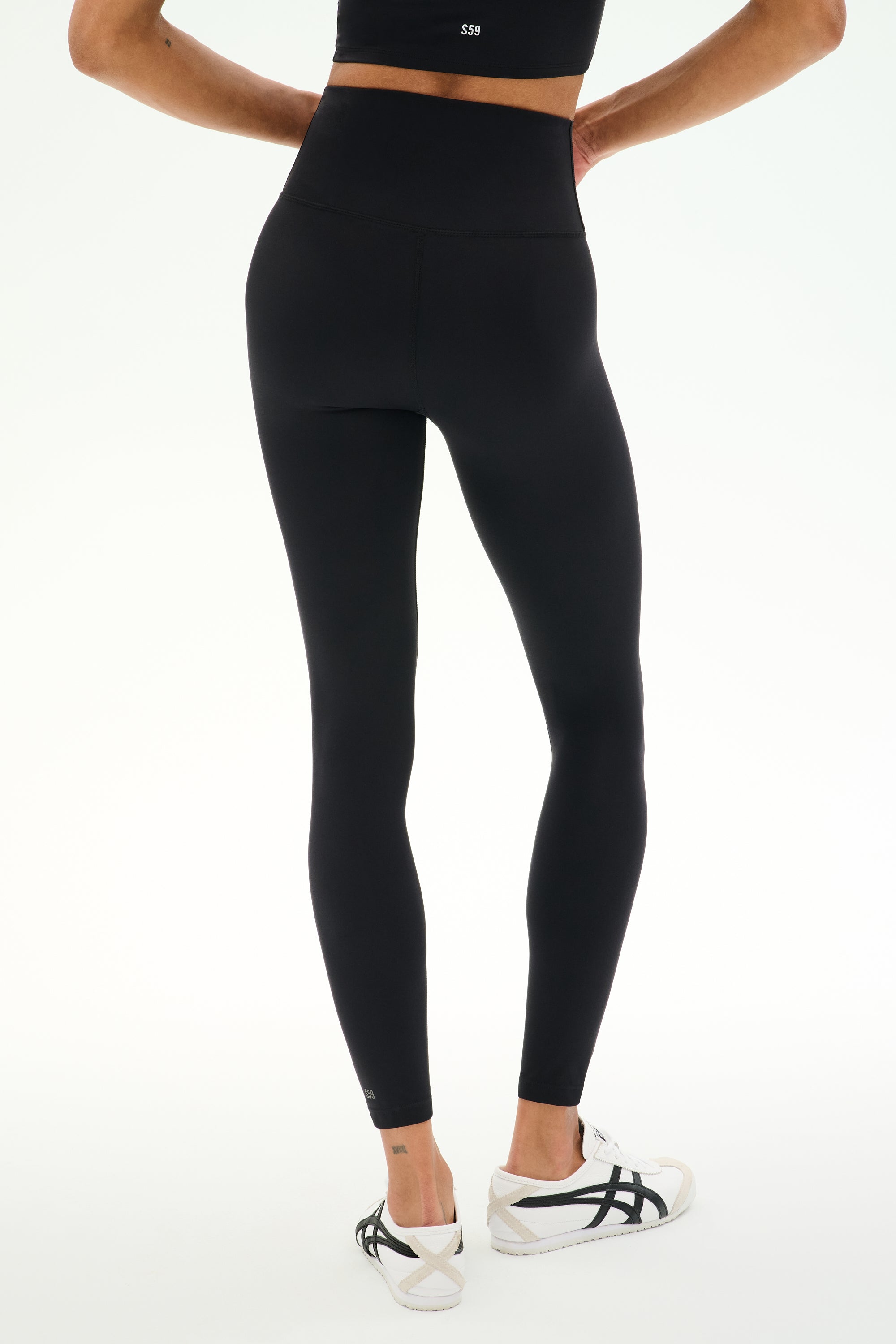 A person is shown from the back standing against a plain white background, wearing SPLITS59 Sprint High Waist Rigor 7/8 leggings in black made from durable Rigor fabric, paired with white athletic shoes.