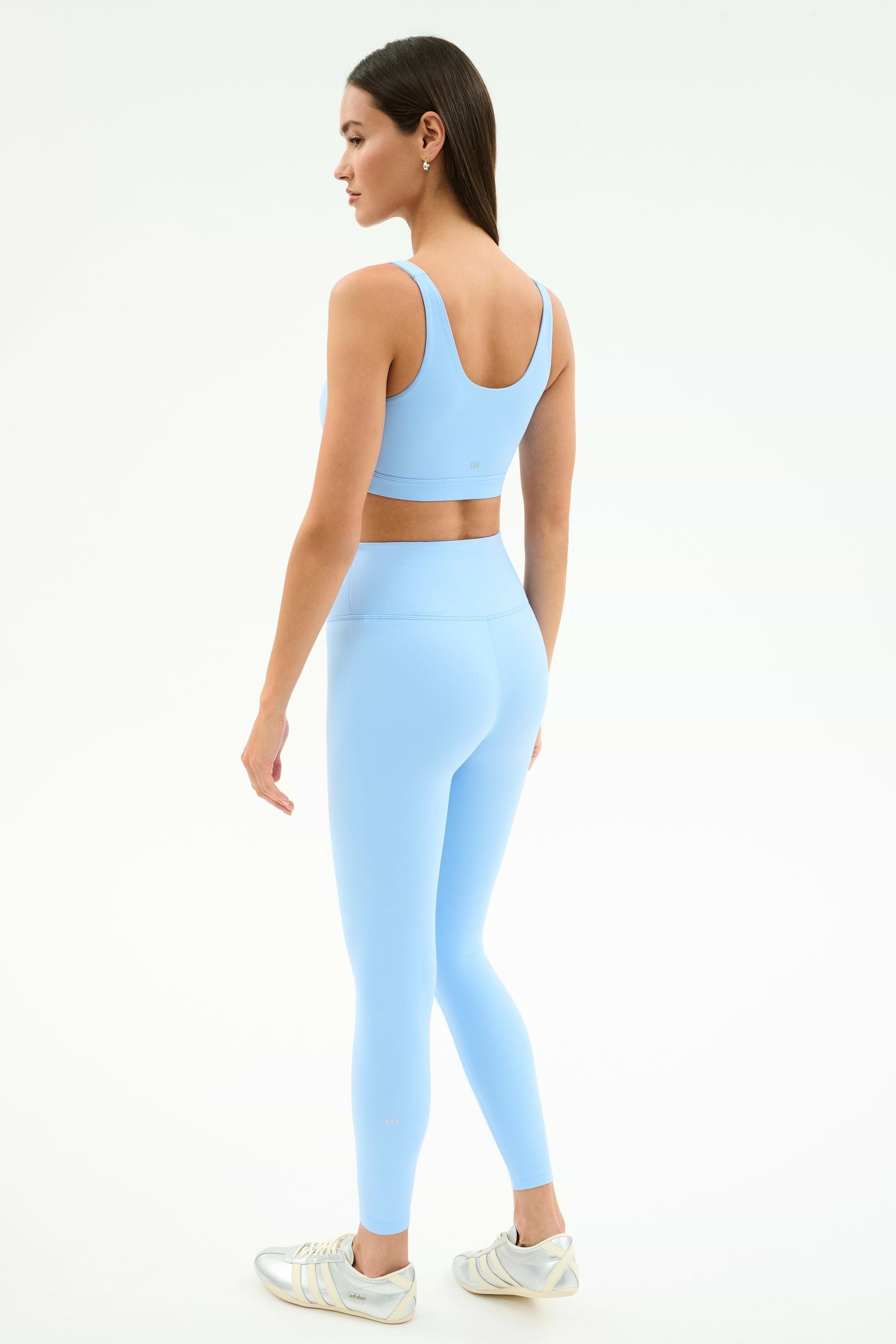 A woman faces away, wearing the SPLITS59 Sprint Rigor Bra in Skylight and matching leggings with white athletic shoes, ready for gym workouts or multi-sport performance against a plain background.