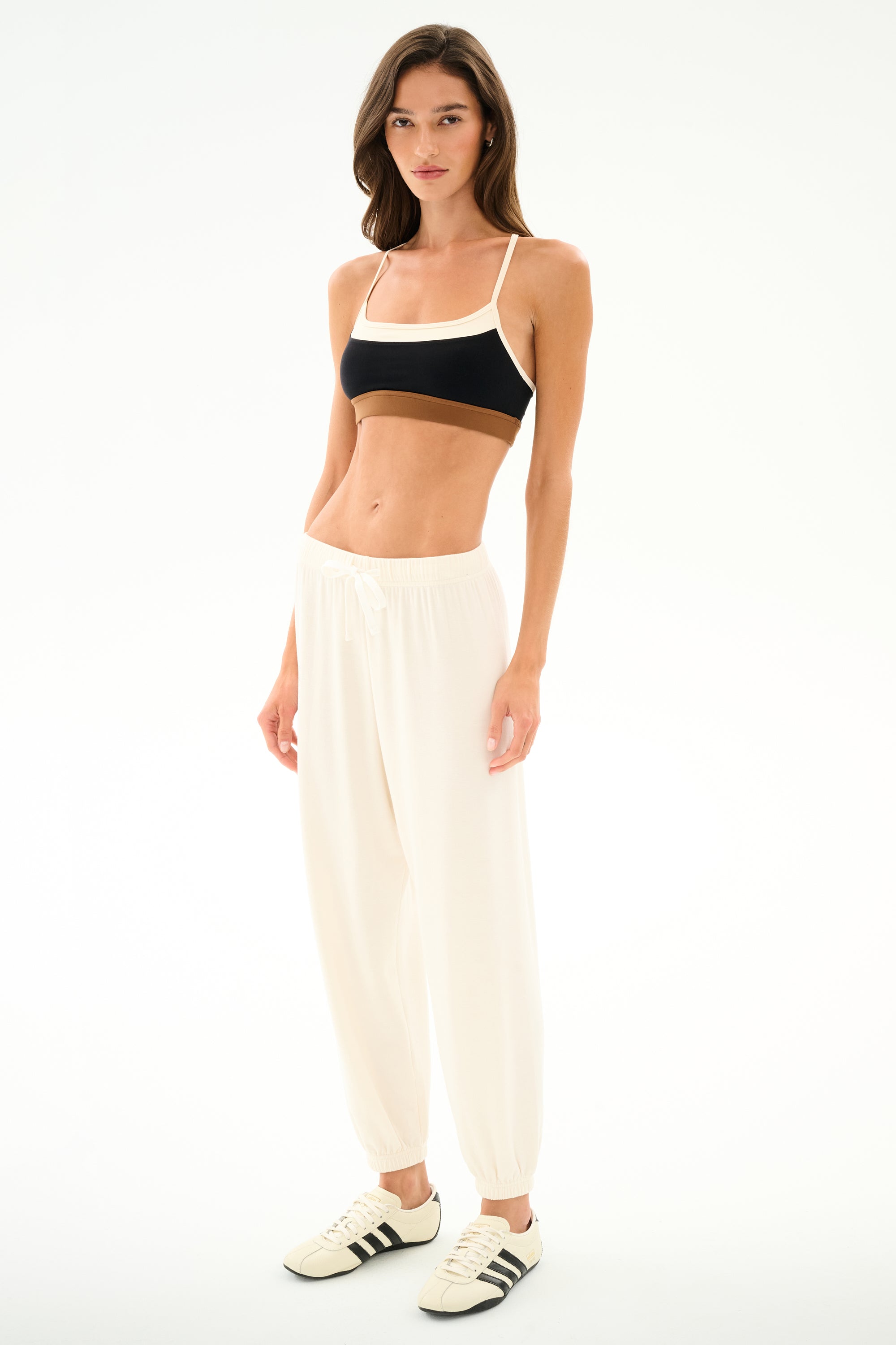 Woman stands on a white background wearing a black sports bra, SPLITS59 Andie Oversized Fleece Sweatpant - Creme, and white sneakers with black stripes.