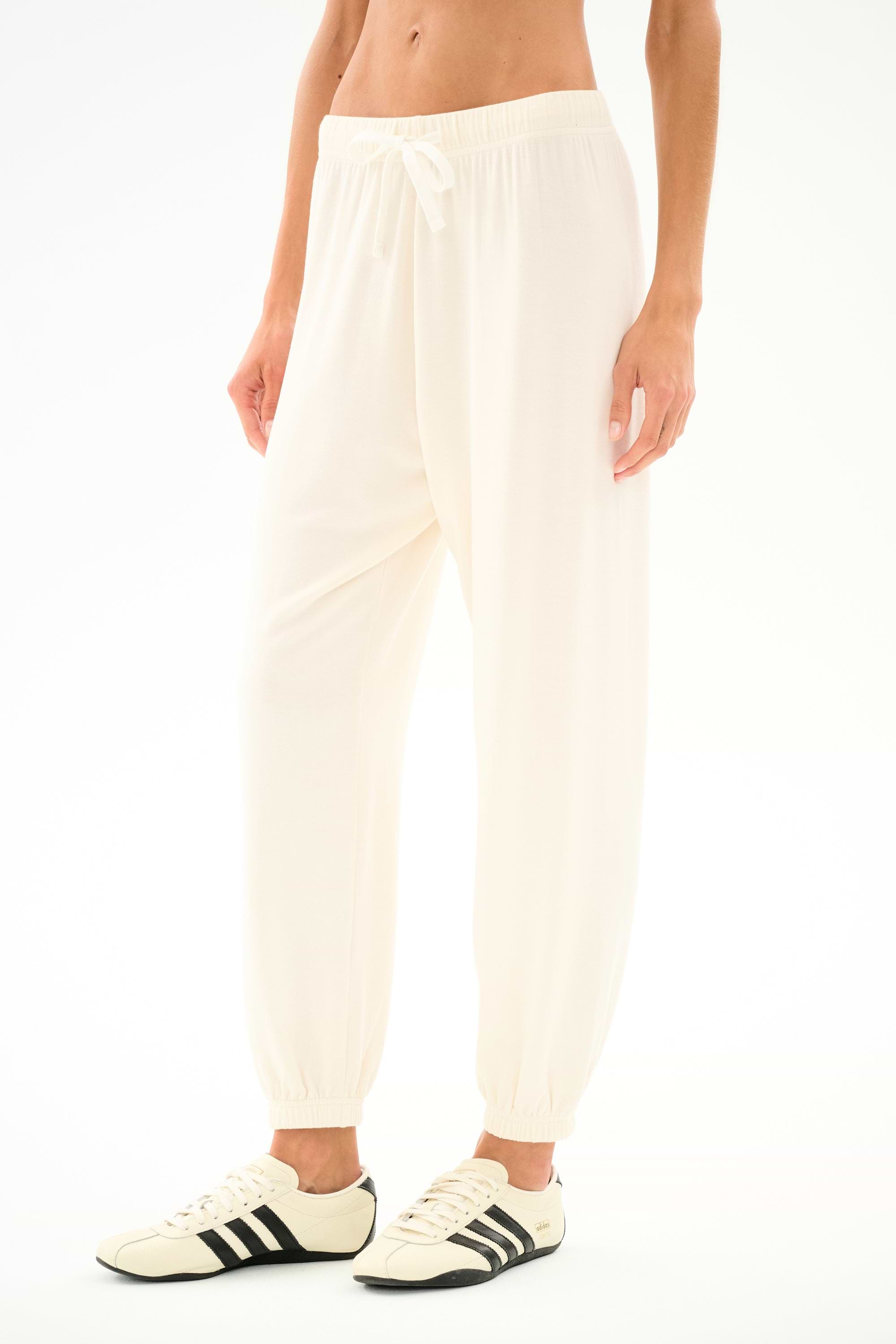 Model wears the SPLITS59 Andie Oversized Fleece Sweatpant - Creme with cream sneakers featuring black stripes, ideal for cool weather workouts, standing against a white backdrop.