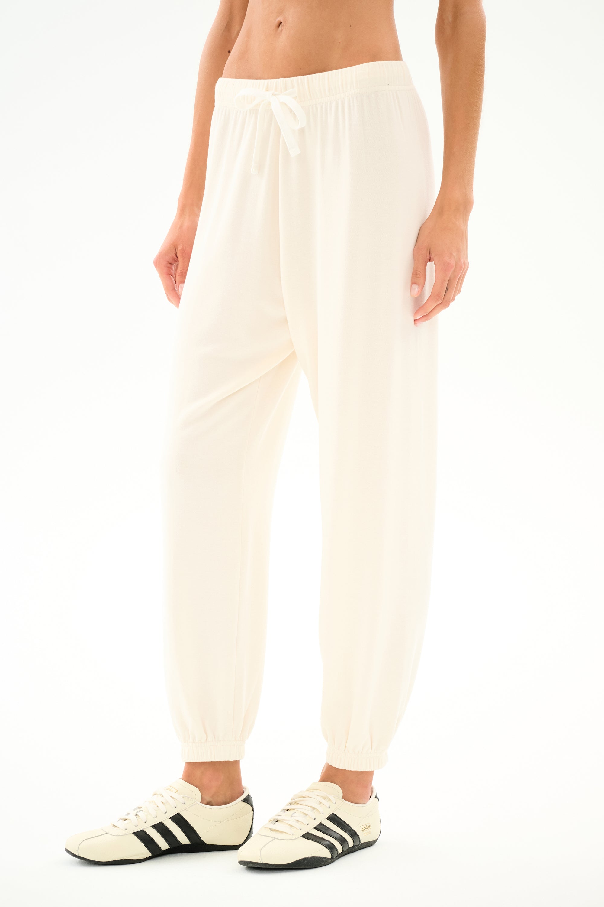Model wears the SPLITS59 Andie Oversized Fleece Sweatpant - Creme with cream sneakers featuring black stripes, ideal for cool weather workouts, standing against a white backdrop.