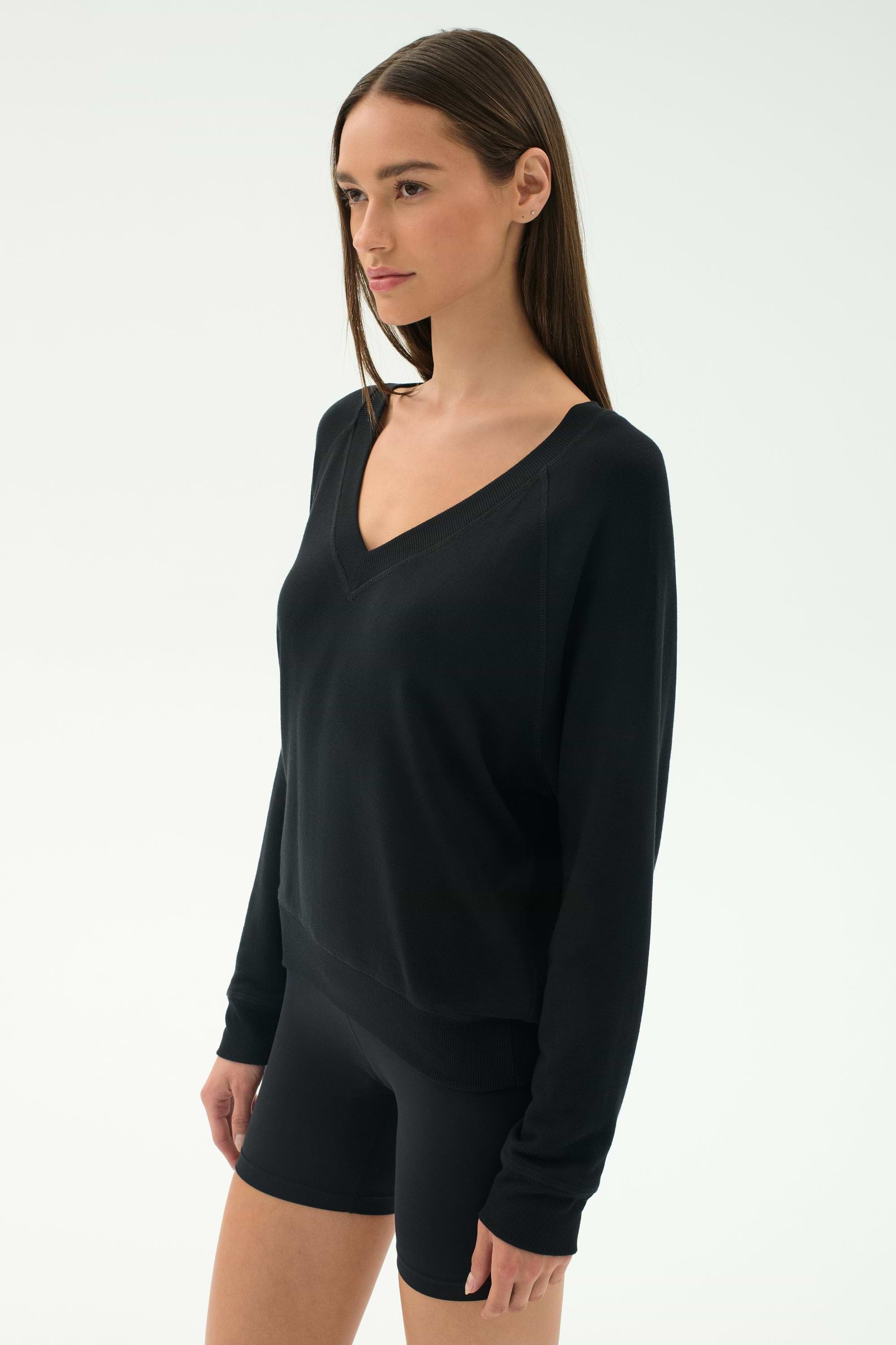 A woman stands against a plain background wearing the SPLITS59 Bennie Fleece V Neck Sweatshirt in black, made in Los Angeles.