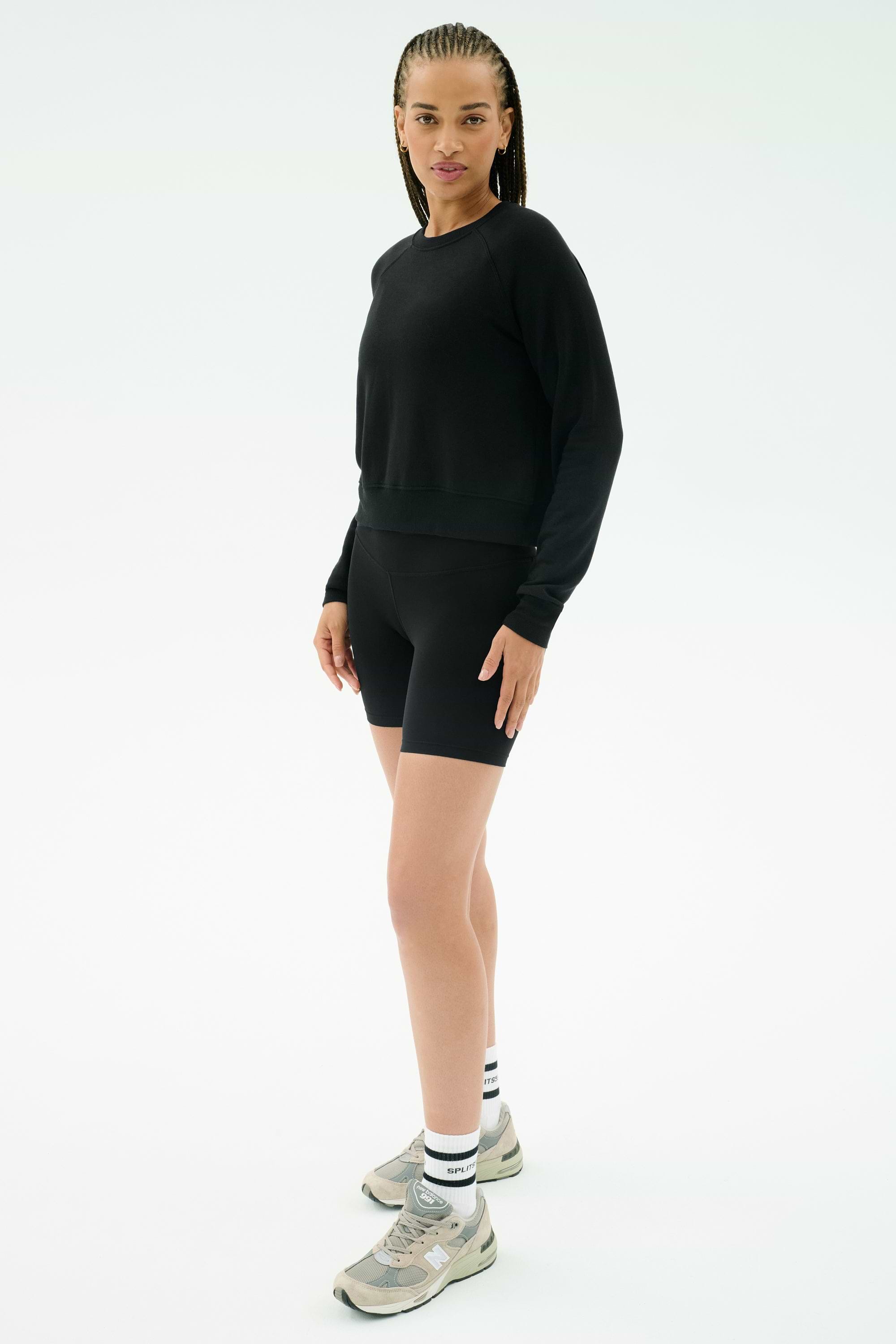A person wearing the SPLITS59 Brooks Fleece Long Sleeve Sweatshirt in black, black bike shorts, white socks, and gray sneakers stands against a plain white background.