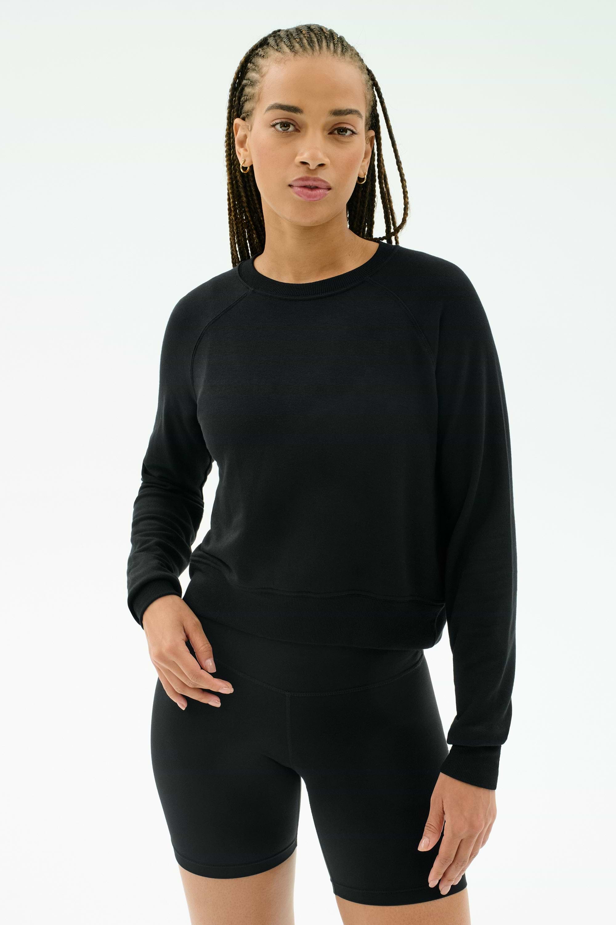 A woman wears the SPLITS59 Brooks Fleece Long Sleeve Sweatshirt in black, paired with fitted shorts, standing against a plain white background. Made in Los Angeles.