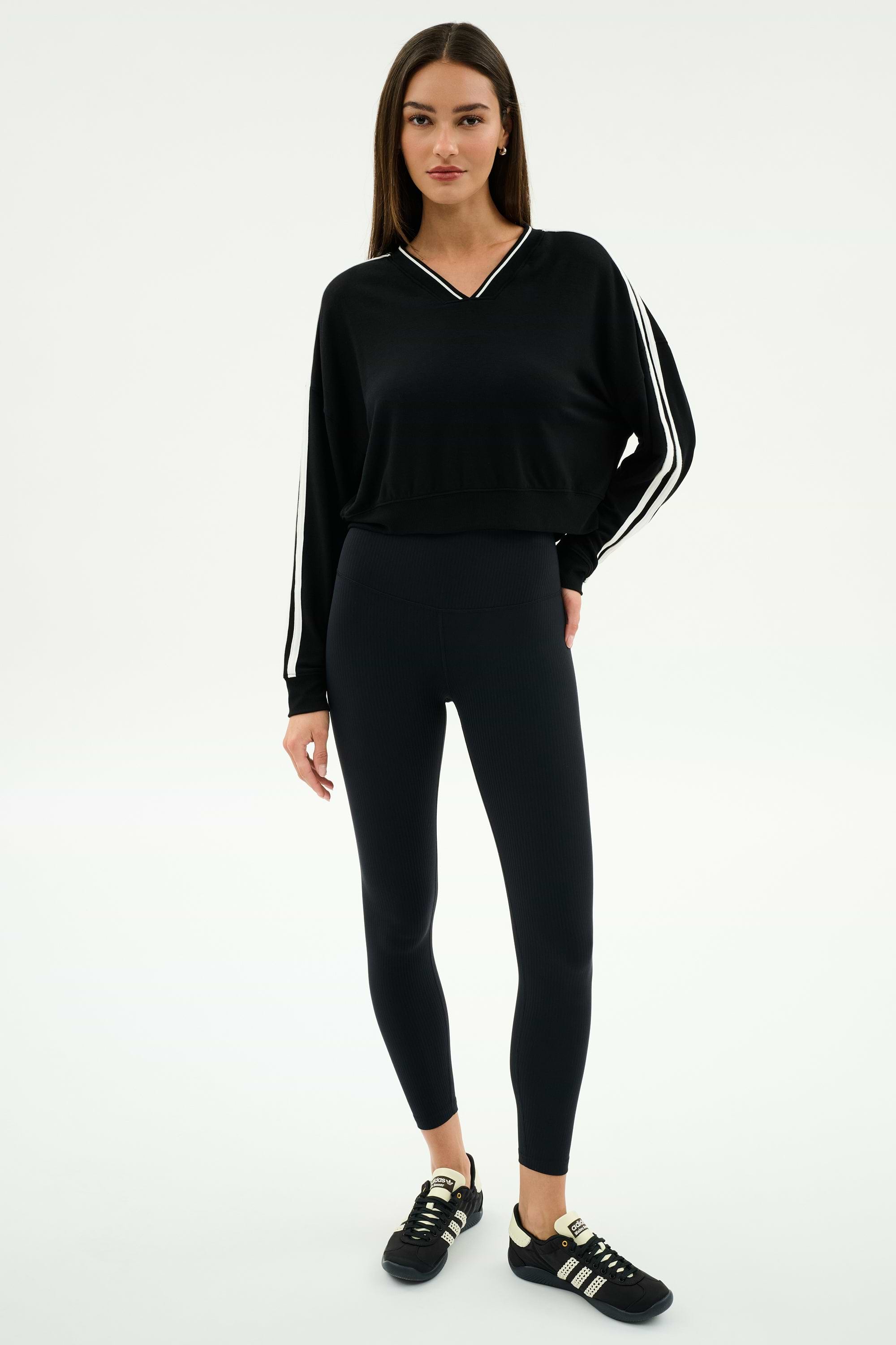A woman wears the SPLITS59 Ellie Fleece V Neck Sweatshirt in Black/White, featuring white stripes, paired with black leggings and black sneakers with white accents, standing against a plain background.