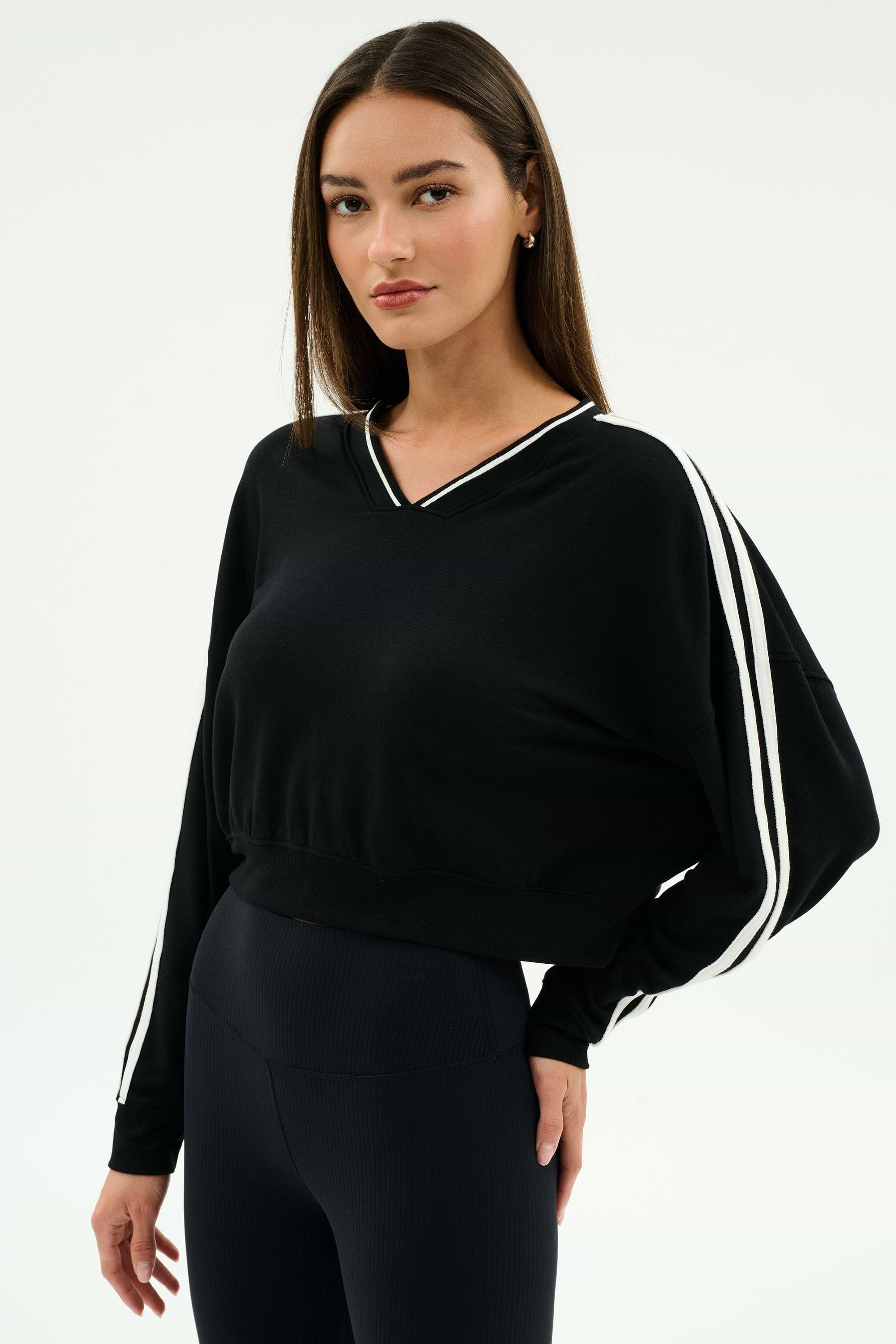A woman stands against a plain background, wearing the SPLITS59 Ellie Fleece V Neck Sweatshirt in Black/White and black leggings designed for comfort and flexibility.