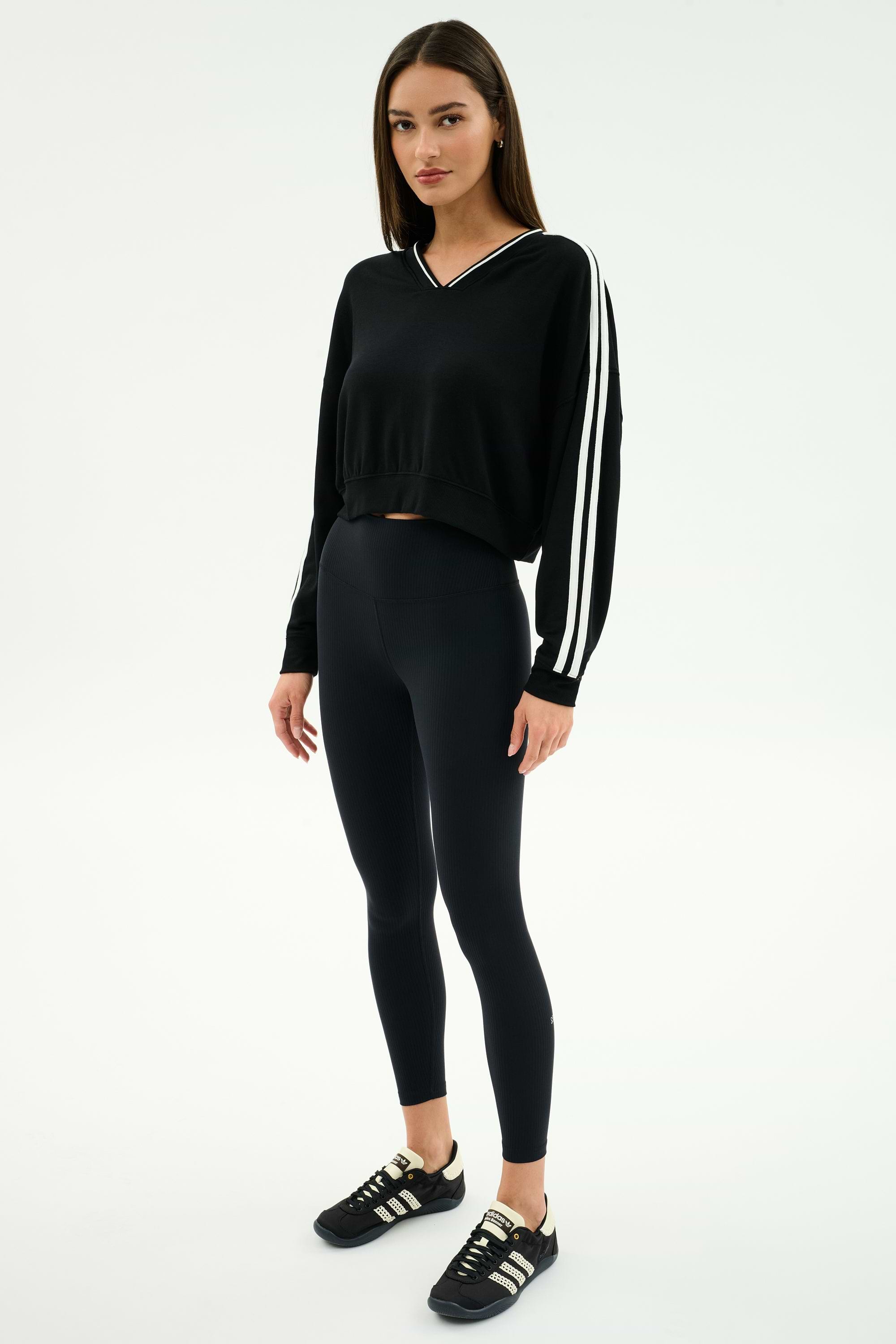 A woman stands in a studio wearing the SPLITS59 Ellie Fleece V Neck Sweatshirt in black/white, paired with black leggings and sneakers.