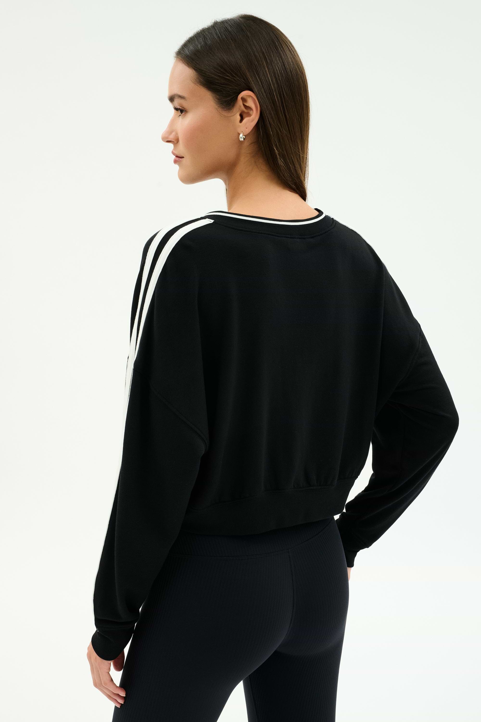 A woman stands with one hand on her hip, modeling the SPLITS59 Ellie Fleece V Neck Sweatshirt in Black/White and black spandex blend leggings, facing away from the camera against a plain background.
