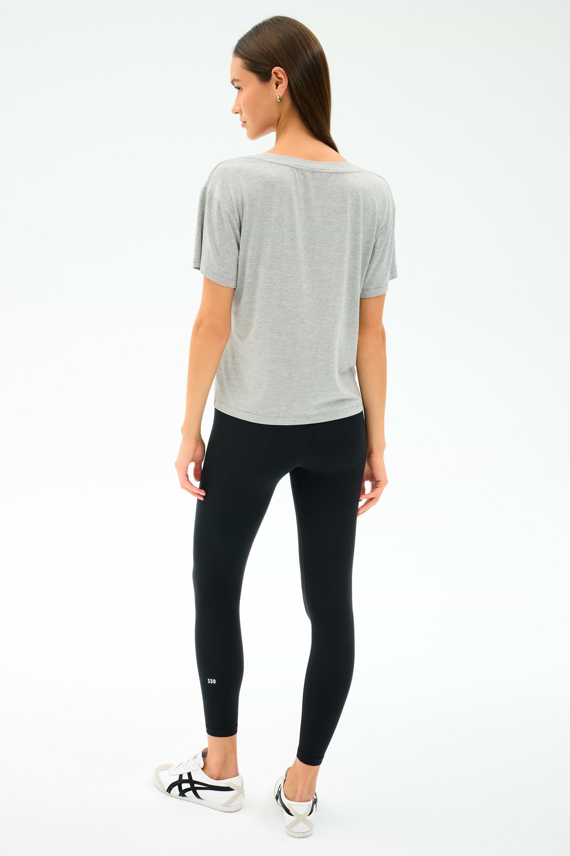 A woman stands facing away from the camera, wearing the SPLITS59 Georgia Jersey V-Neck in Heather Grey, black spandex leggings, and white sneakers against a plain white background.