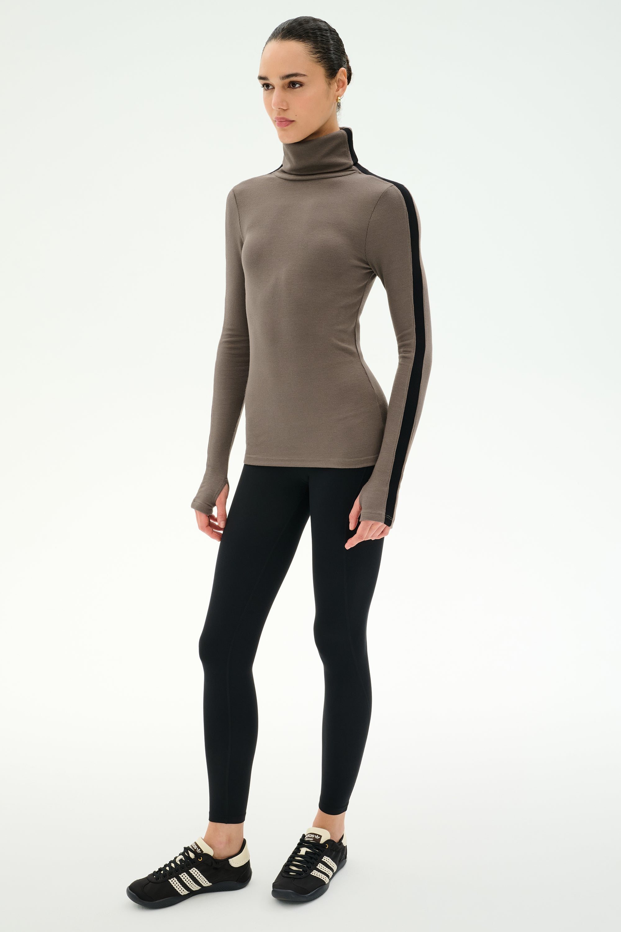 A woman stands against a white background wearing the SPLITS59 Jackson Rib Full Length Turtleneck in Lentil/Black—a soft modal spandex blend top with black striped sleeves—paired with black leggings and black sneakers.