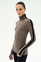 A person wearing the SPLITS59 Jackson Rib Full Length Turtleneck in Lentil/Black—a taupe turtleneck top crafted from a soft modal spandex blend with a black stripe down the sleeve—paired with black pants, stands against a plain white background.