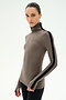 A person wearing the SPLITS59 Jackson Rib Full Length Turtleneck in Lentil/Black—a taupe turtleneck top crafted from a soft modal spandex blend with a black stripe down the sleeve—paired with black pants, stands against a plain white background.