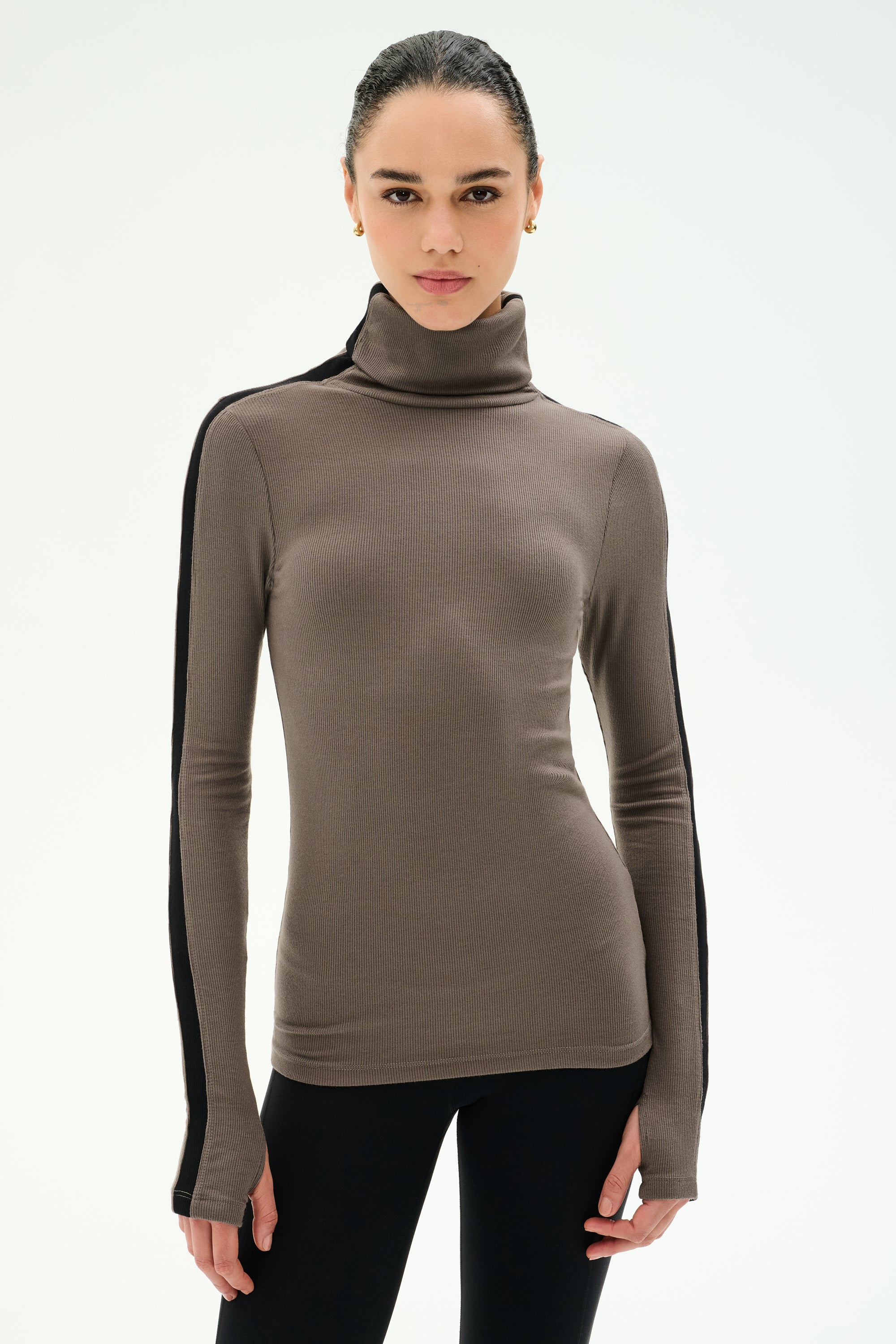 A woman wears the SPLITS59 Jackson Rib Full Length Turtleneck in Lentil/Black—a brown ribbed modal spandex top with black striped sleeves—paired with black leggings and standing against a plain white background.