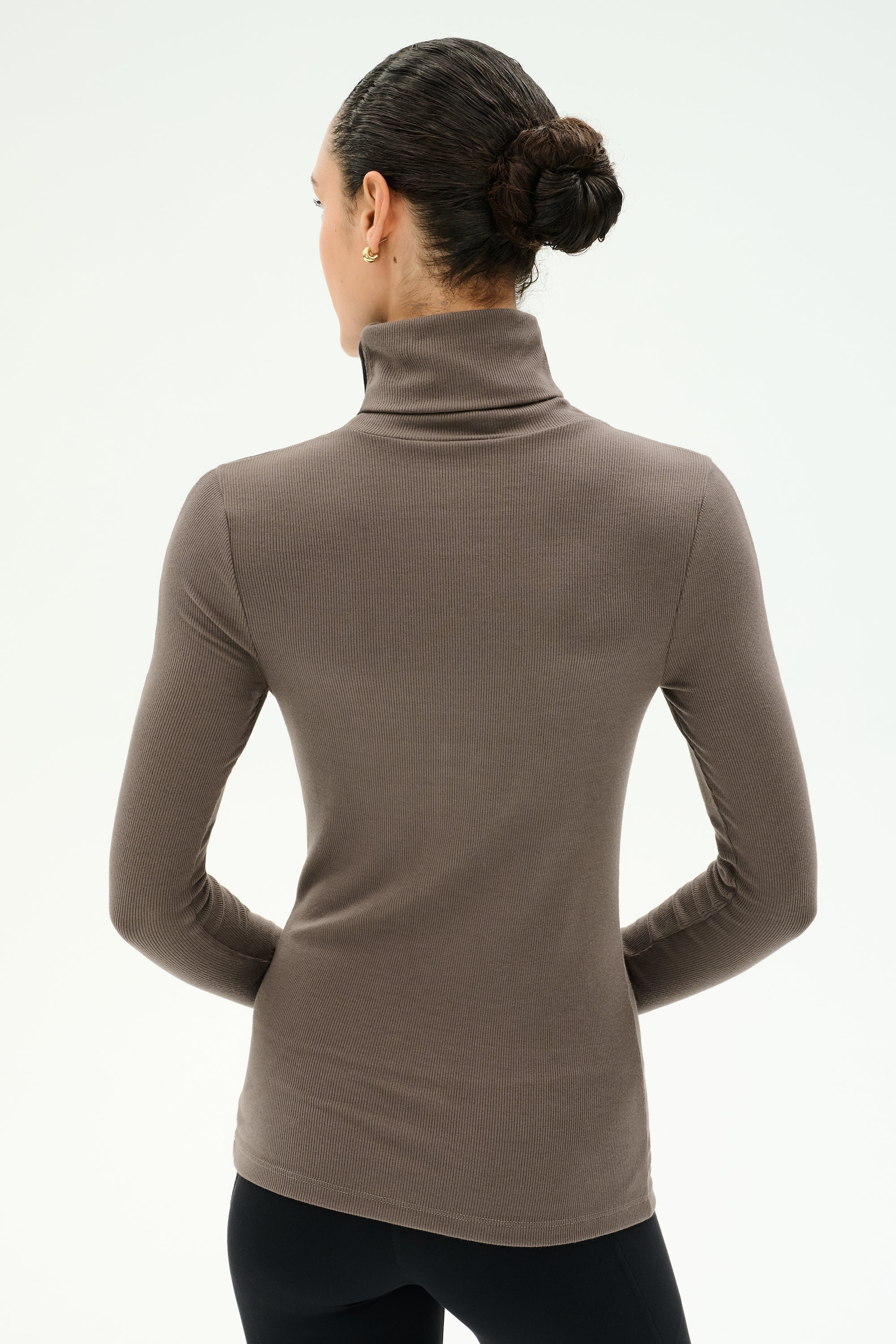 A person with dark hair in a bun wears the SPLITS59 Jackson Rib Full Length Turtleneck in Lentil, paired with black pants, standing with their back to the camera against a light background.