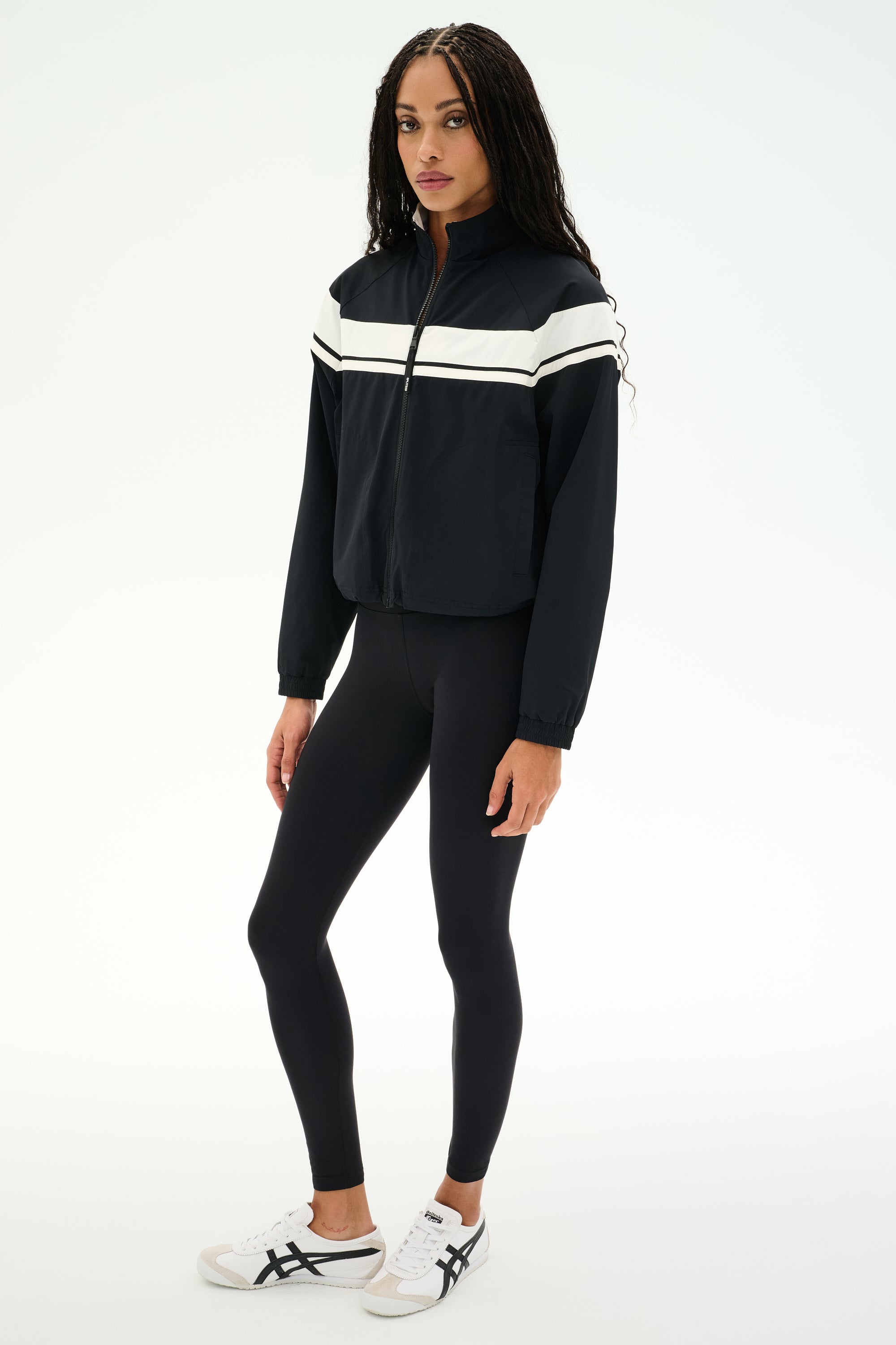 A person is wearing the SPLITS59 Morrison Nylon Twill Track Jacket in Black/Creme, paired with black spandex leggings and white sneakers, standing against a plain white background.