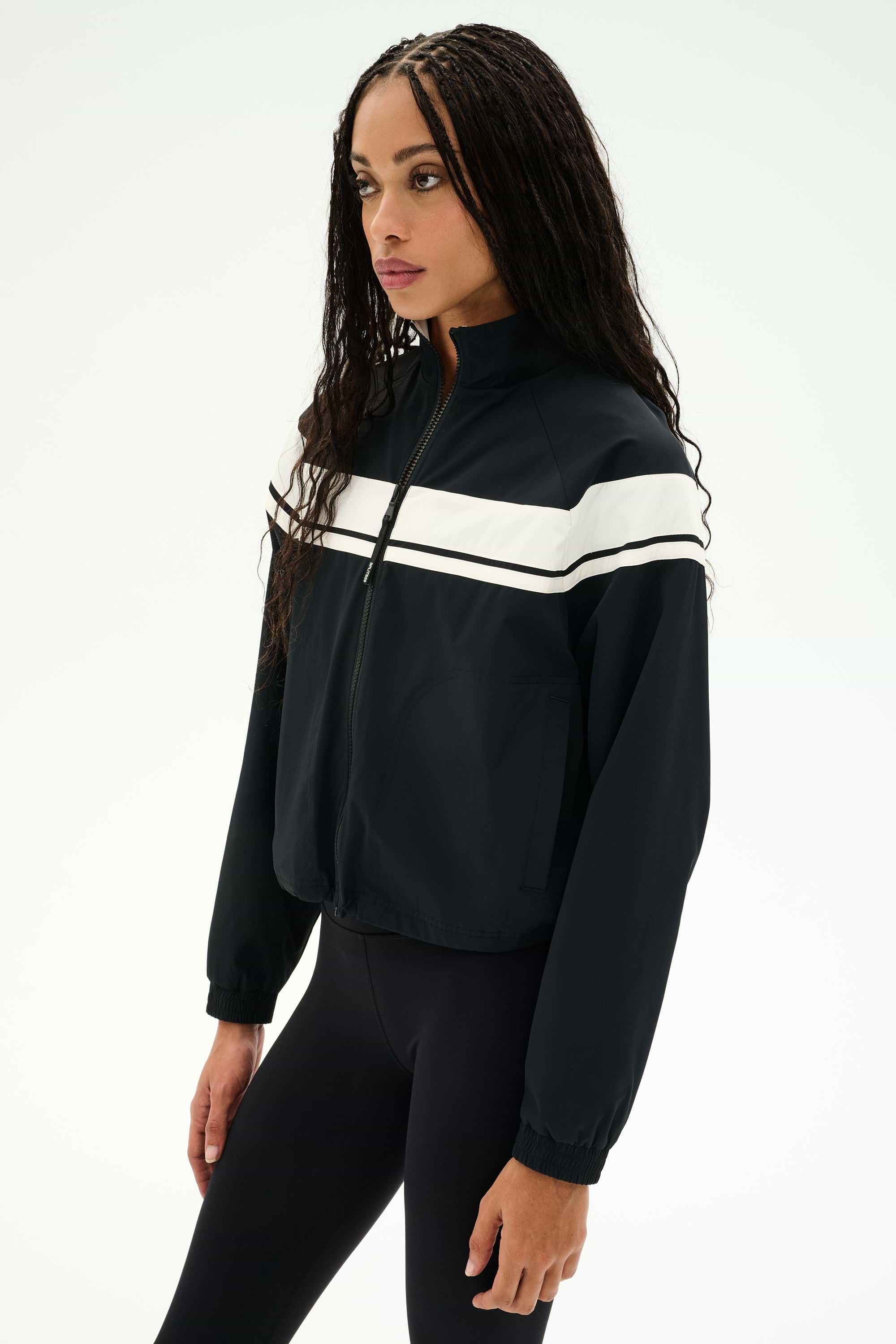 A person is wearing the SPLITS59 Morrison Nylon Twill Track Jacket in Black/Creme and black spandex leggings, standing against a plain white background.