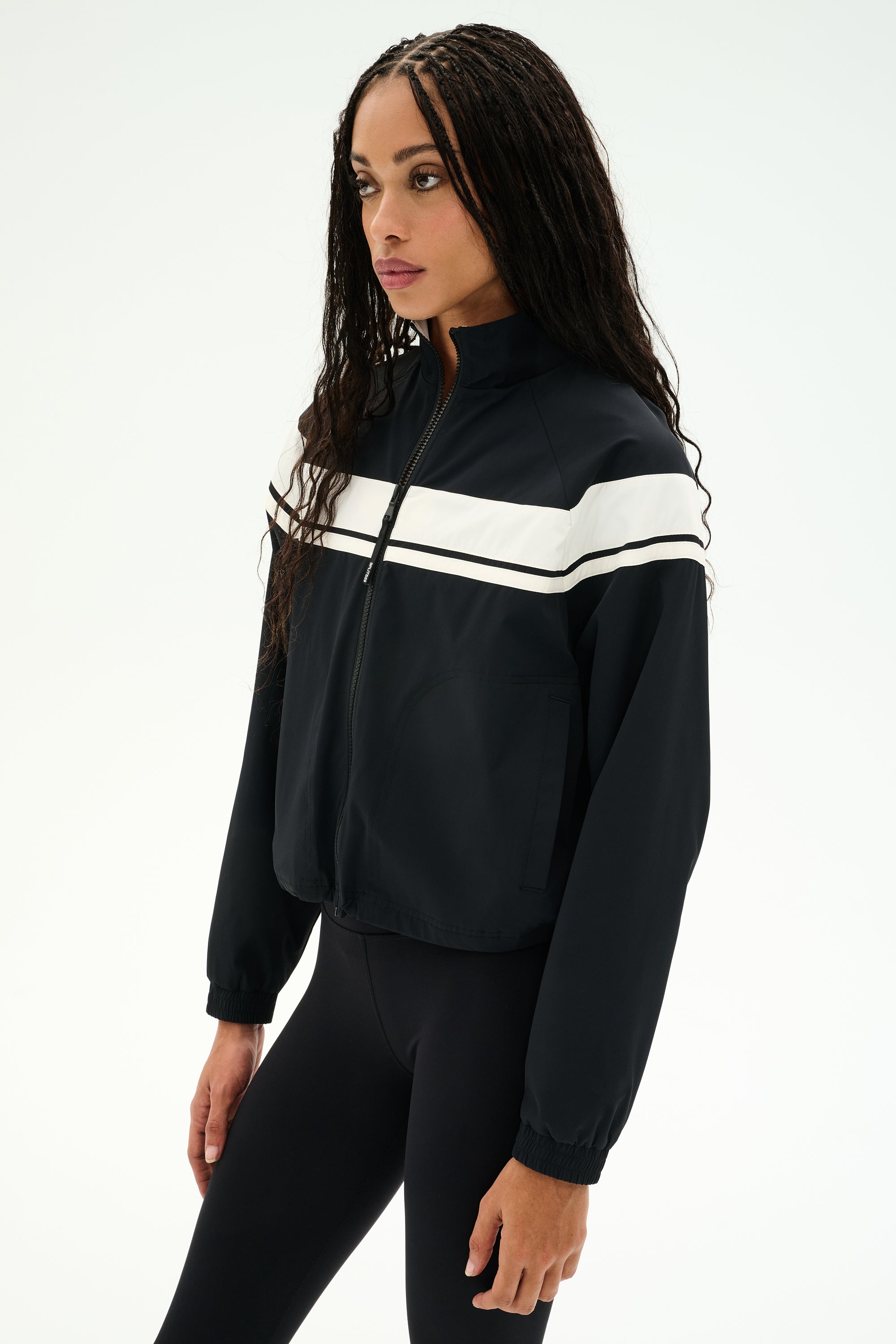 A person is wearing the SPLITS59 Morrison Nylon Twill Track Jacket in Black/Creme and black spandex leggings, standing against a plain white background.