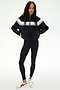 A woman stands against a white background wearing the SPLITS59 Morrison Nylon Twill Track Jacket in Black/Creme, paired with black spandex leggings and white sneakers with black stripes.