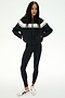 A woman stands against a white background wearing the SPLITS59 Morrison Nylon Twill Track Jacket in Black/Creme, paired with black spandex leggings and white sneakers with black stripes.