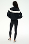 A person stands with their back to the camera against a plain white background, wearing the SPLITS59 Morrison Nylon Twill Track Jacket in Black/Creme, paired with black spandex leggings and white sneakers.