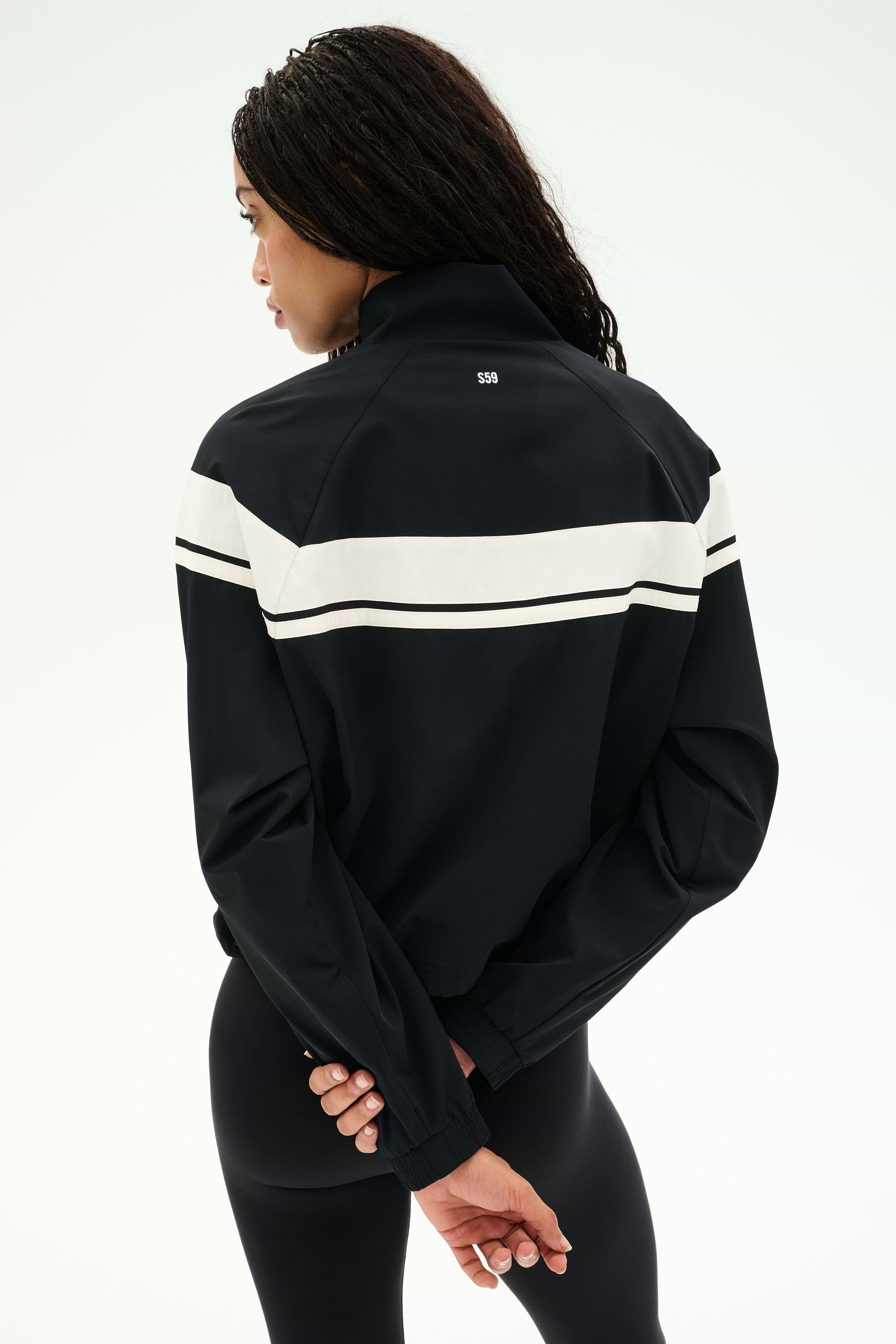A person stands with their back to the camera, wearing the SPLITS59 Morrison Nylon Twill Track Jacket in Black/Creme, featuring a white stripe across the back, paired with black spandex leggings. Their hands are clasped behind their back.
