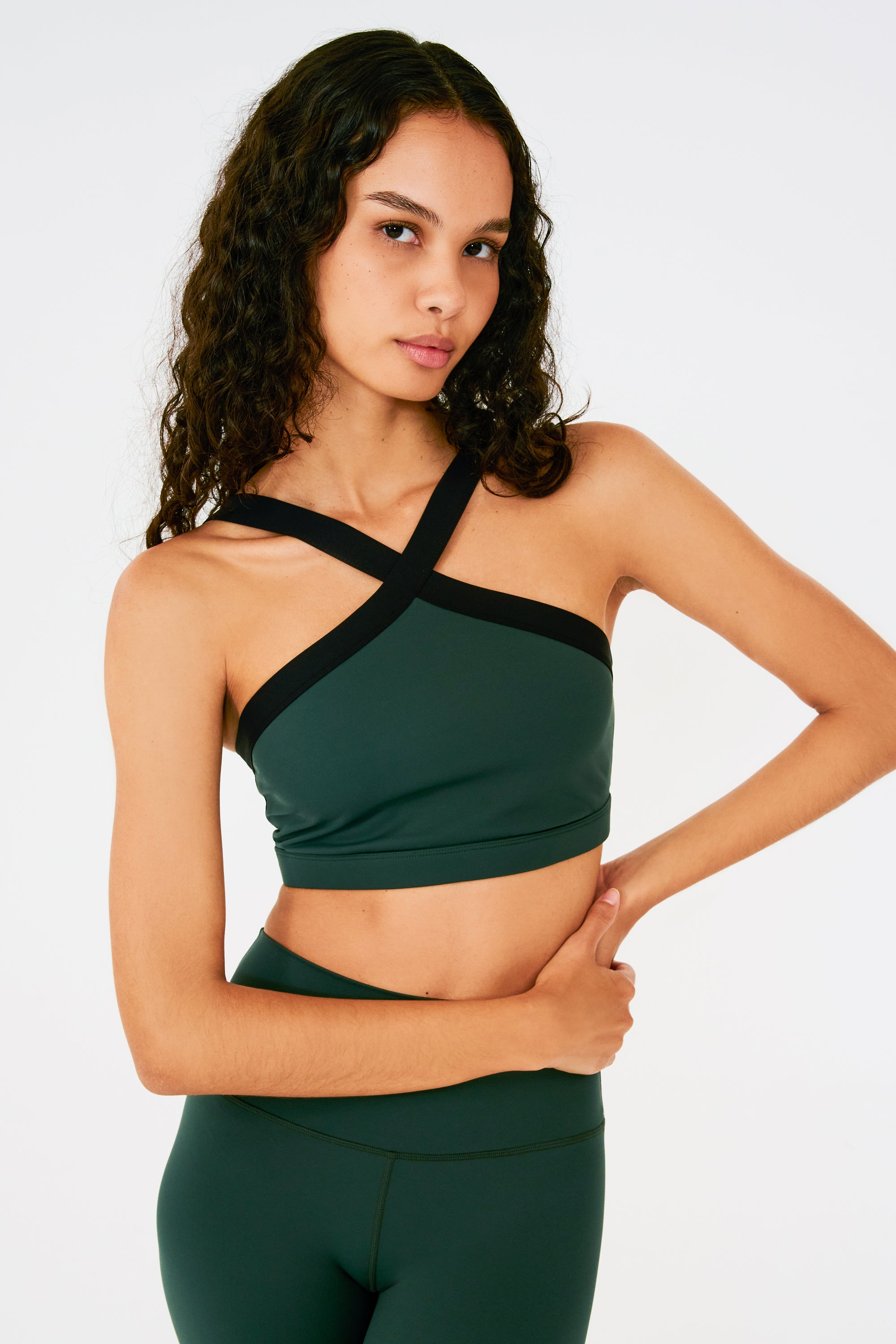 Front view of woman with dark wavy hair wearing a dark green bra with thick black cross-front neckline straps and dark green leggings.