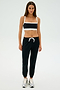 A person stands against a plain backdrop, dressed in a black and white sports bra, SPLITS59's Sonja Fleece Sweatpant in Black suited for cool weather workouts, and silver sneakers.
