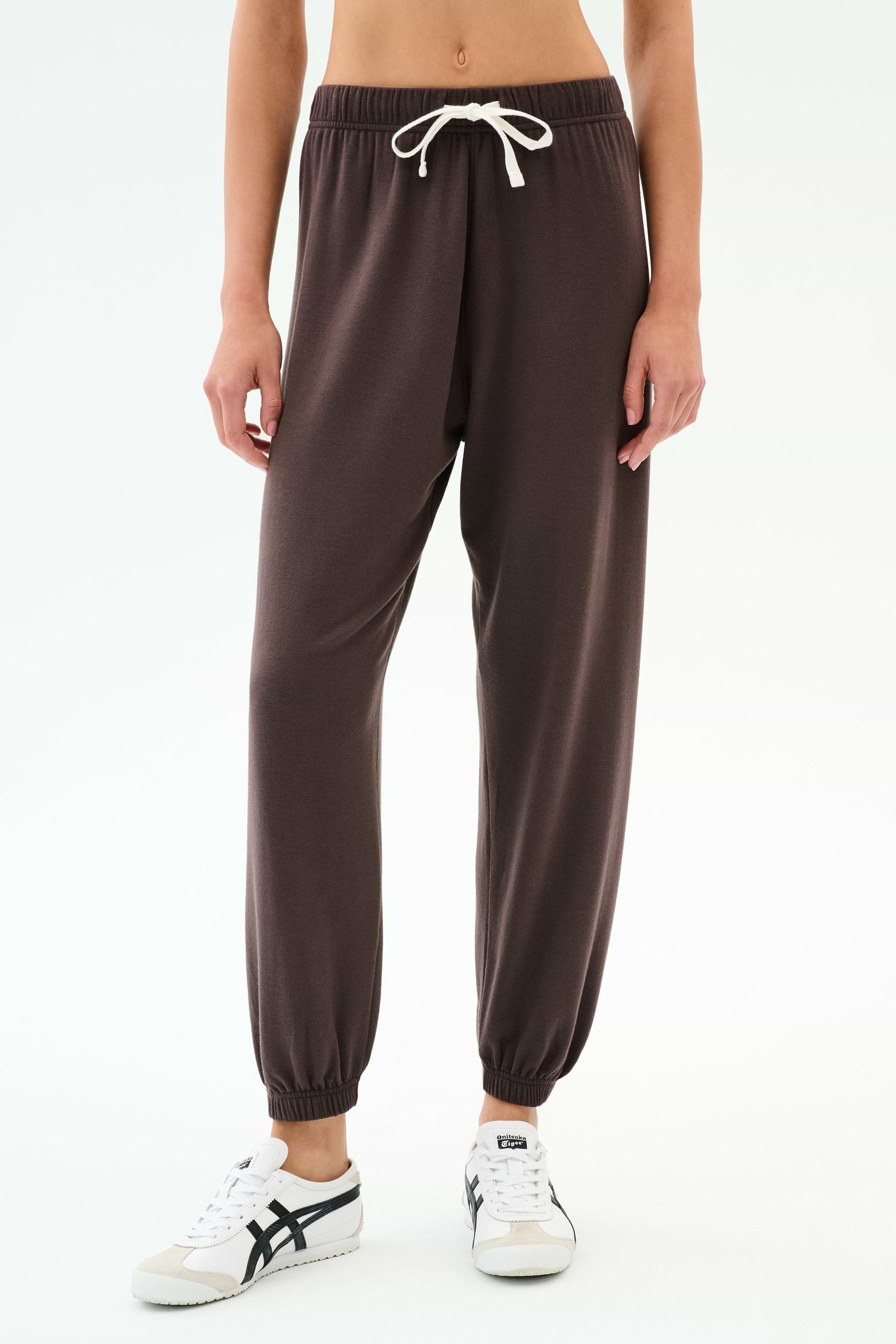 A person wears SPLITS59's Andie Oversized Fleece Sweatpant in Dark Chocolate with a white drawstring and white sneakers, standing against a plain white background. These ultra luxe fleece sweatpants are made in Los Angeles.