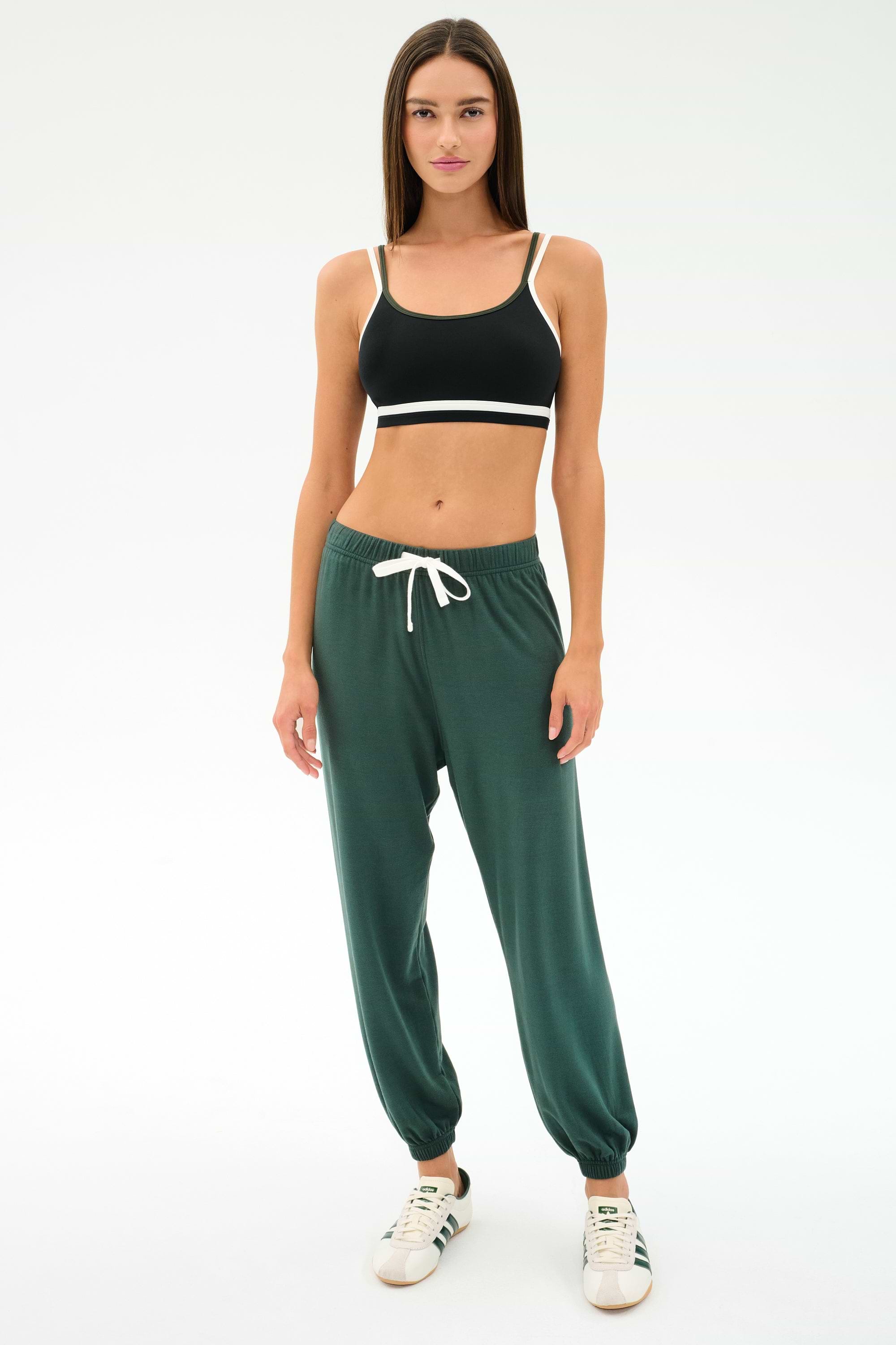 A woman faces forward in a black sports bra, SPLITS59's Andie Oversized Fleece Sweatpant in Military, and white sneakers—ideal for cool weather workouts—set against a plain white background.