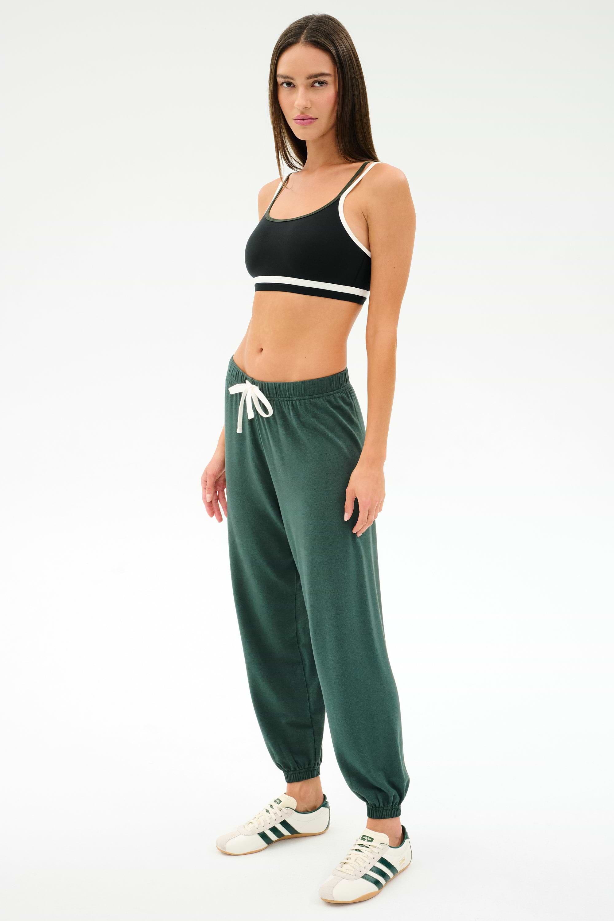 A woman stands against a plain background in a black sports bra, SPLITS59 Andie Oversized Fleece Sweatpant in Military, and white sneakers with green stripes—ideal for warm weather workouts.