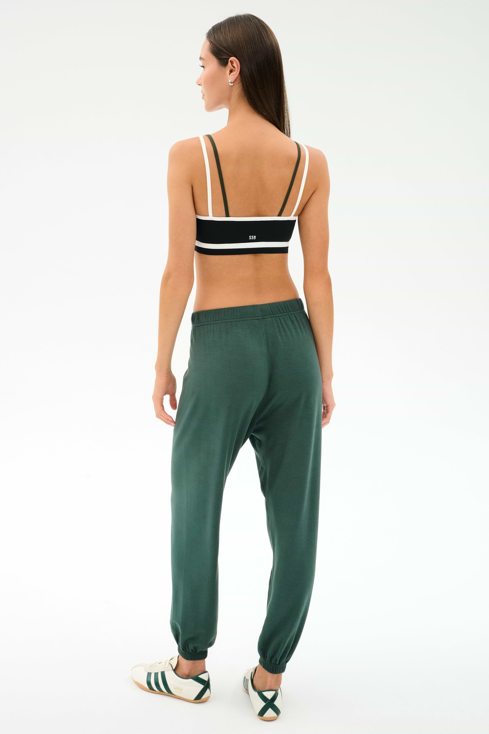 A woman faces away, wearing a black and white sports bra and SPLITS59 Andie Oversized Fleece Sweatpant in Military, paired with white sneakers with green stripes—ideal for cool weather workouts—on a plain light background.