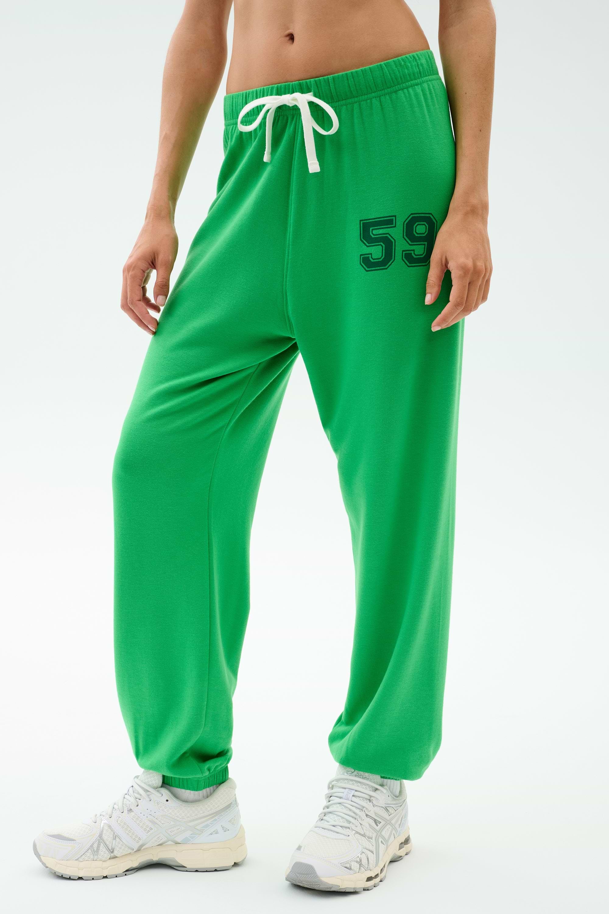 A person wears the SPLITS59 Andie Oversized Fleece Varsity 59 Sweatpant in Grass/59, featuring a bright green color, white drawstring, and number 59 on the left thigh with white athletic shoes against a plain background.