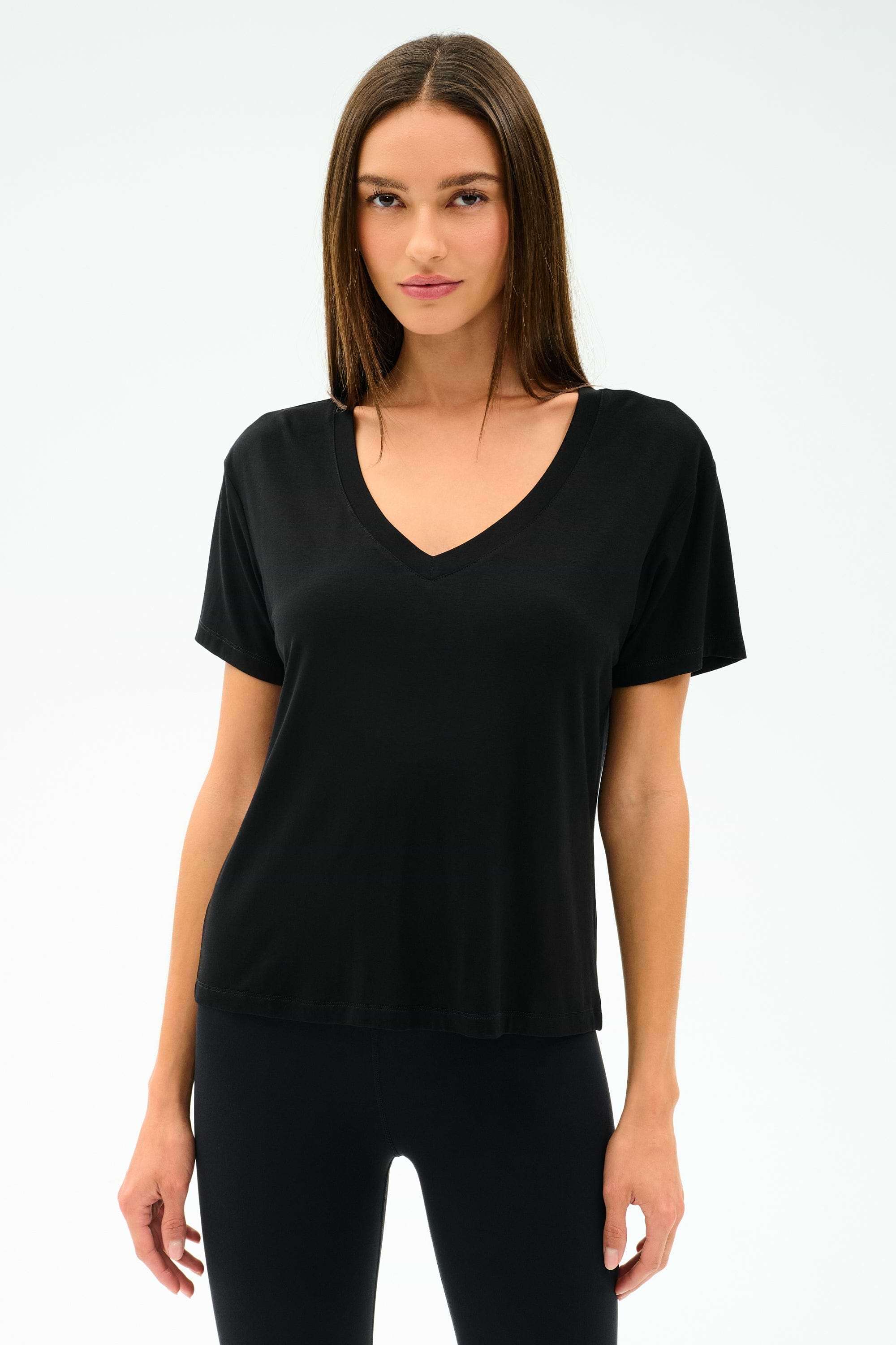A woman faces forward, wearing the SPLITS59 Georgia Jersey V-Neck in black—crafted from a soft viscose-spandex blend—paired with black pants, set against a white background.